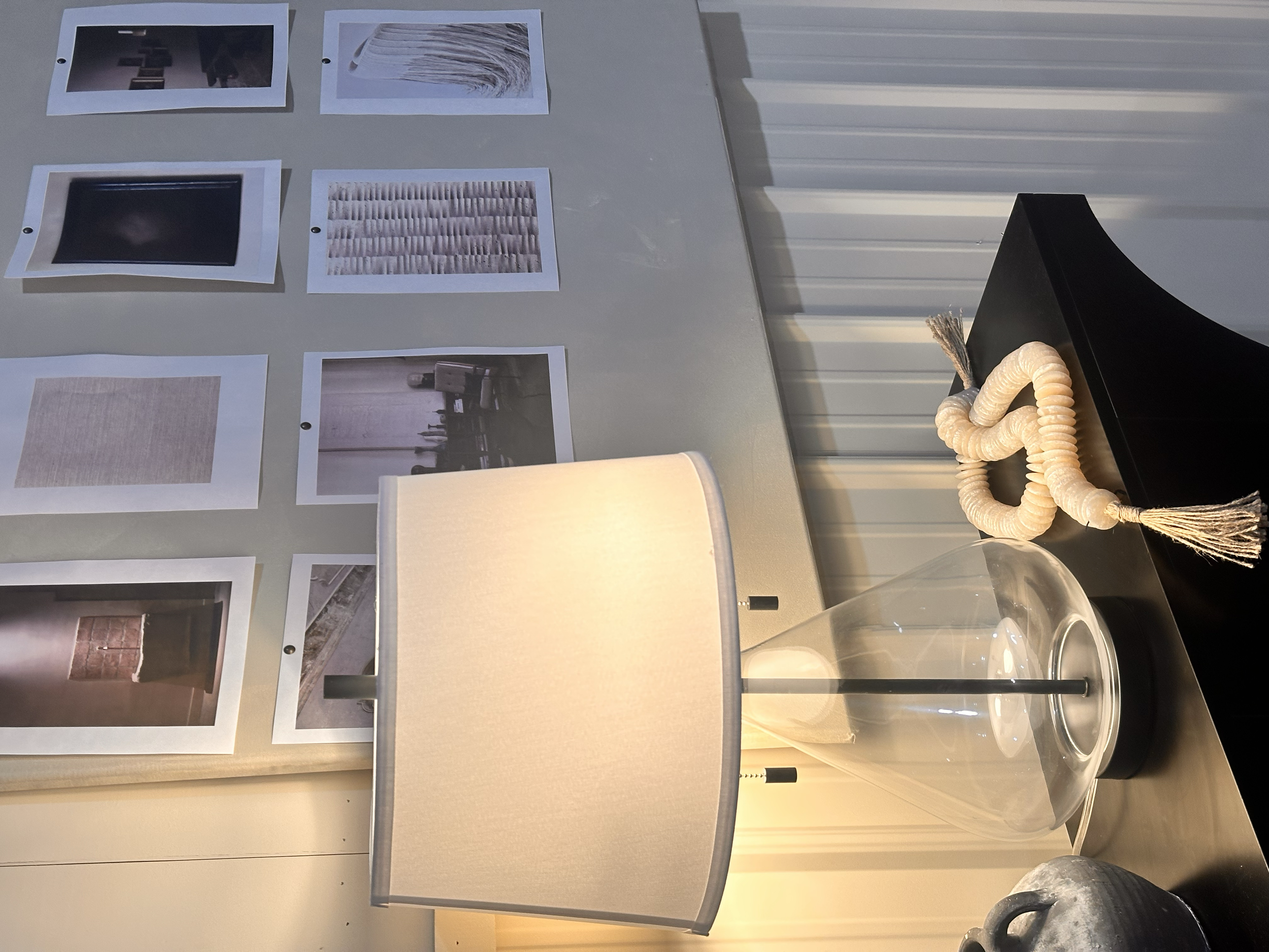 A black table with a large glass vase, a white lamp, and a decorative rope with tassels. Behind the table, there is a wall with nine printed photos arranged in a 3x3 grid.