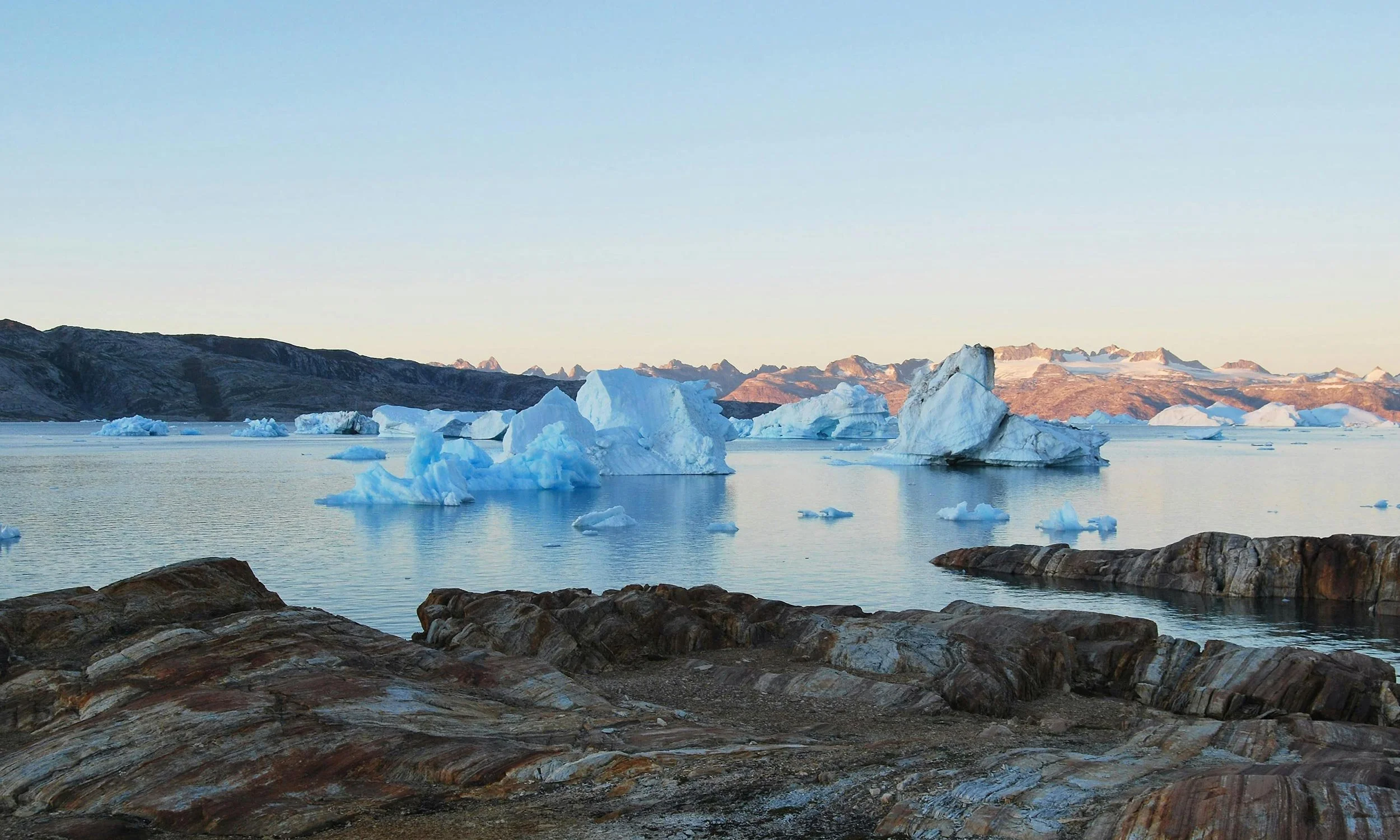 Icebergs floating in calm water with rocky shoreline in foreground, mountains in background, during sunset or sunrise.