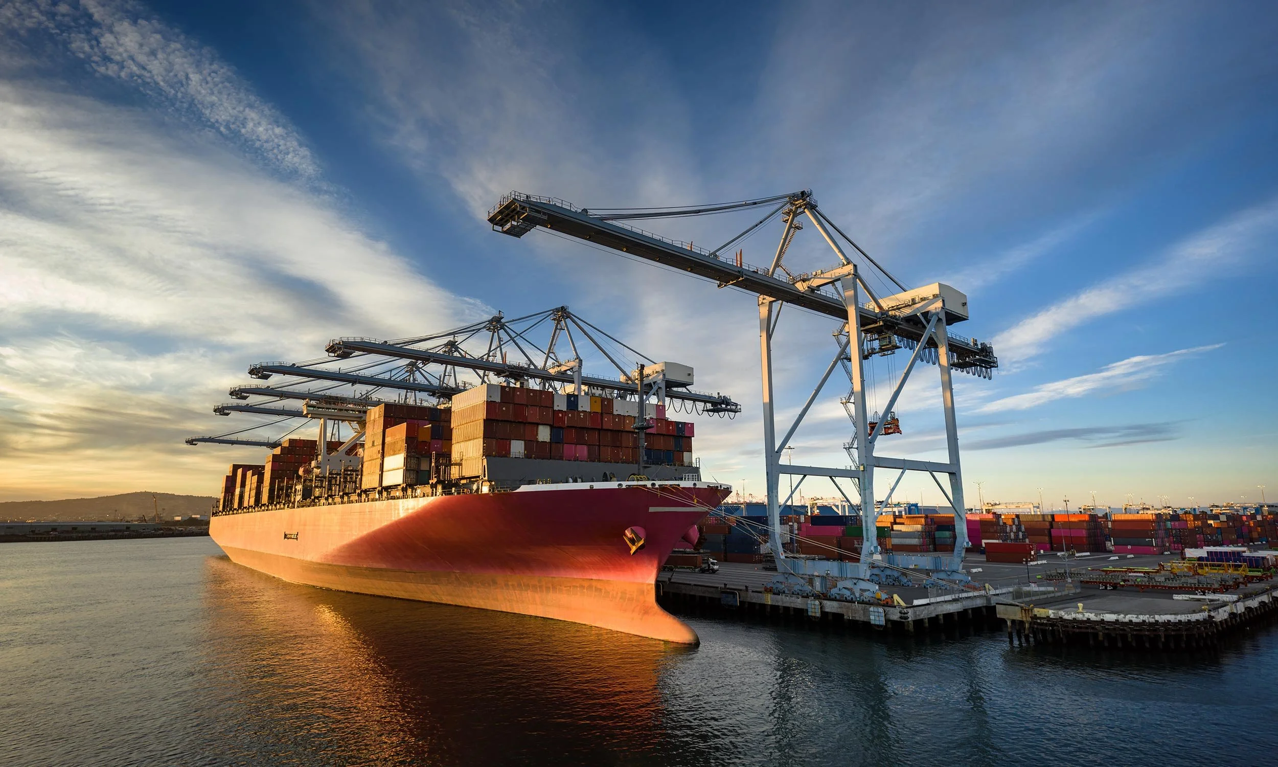 A large cargo ship docked at a port with multiple cranes and containers, under a partly cloudy sky during sunset.