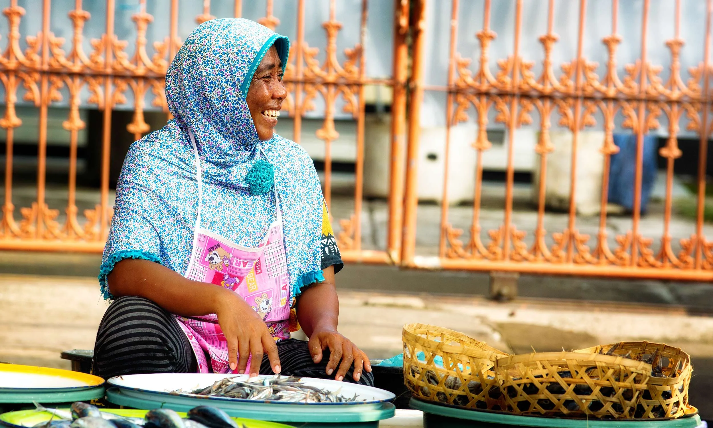 A woman wearing a patterned blue headscarf and apron smiling while selling fish at an outdoor market, with fish displayed on green trays and a woven basket in front of her.
