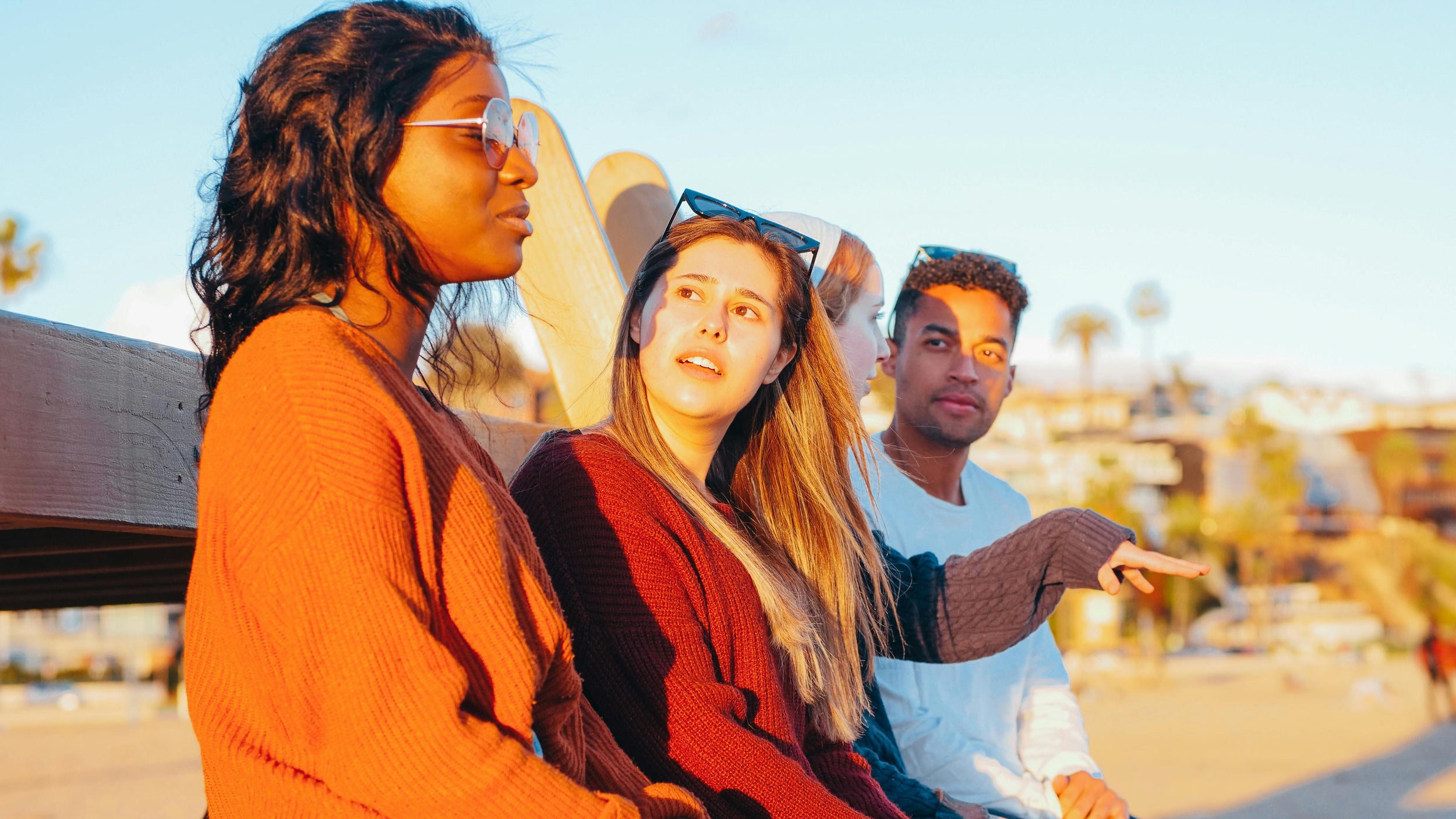 Four diverse adults sitting on a bench outdoors by the sea during sunset, engaged in conversation.