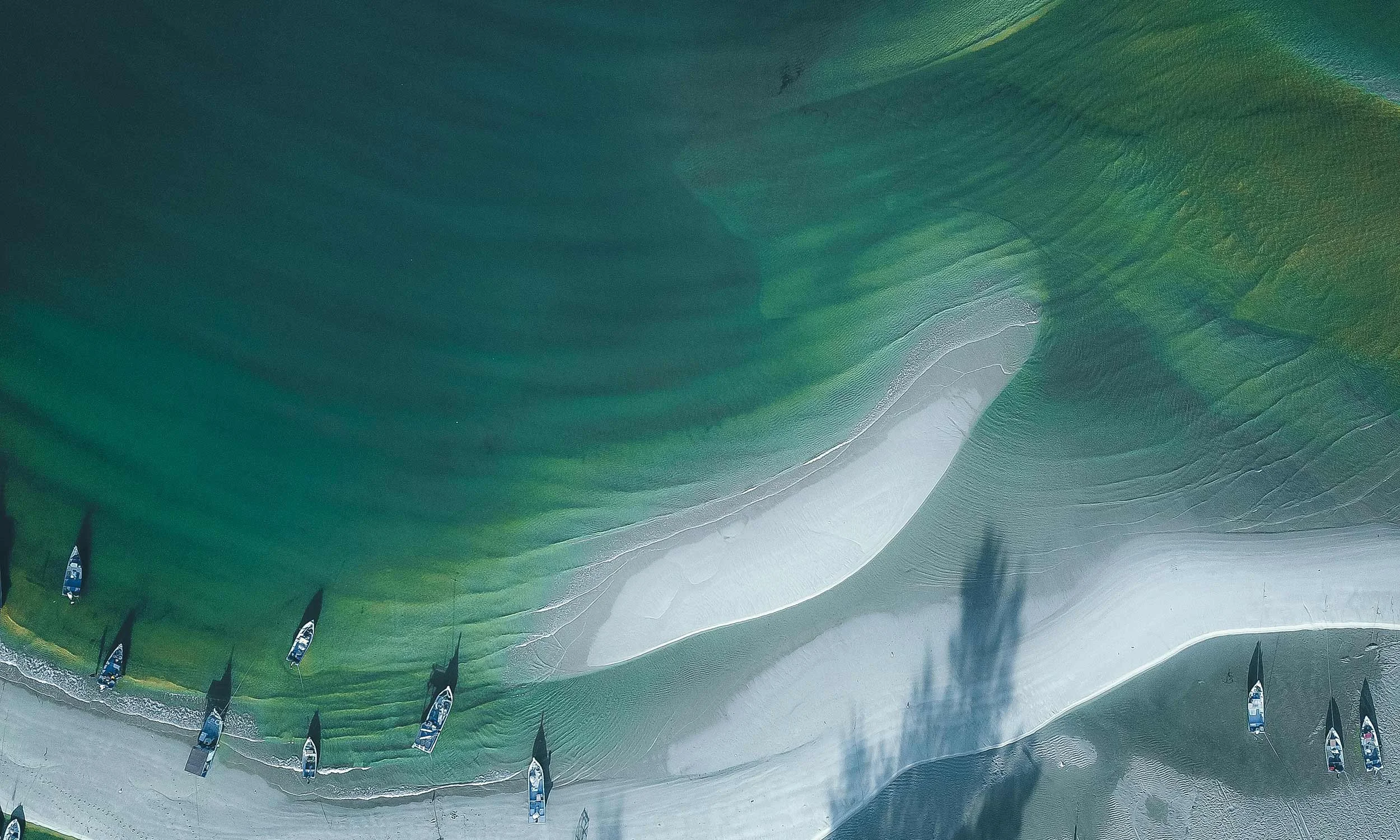 Aerial shot of a sandy coastline with clear green water and artisanal fishing vessels pulled up on the foreshore
