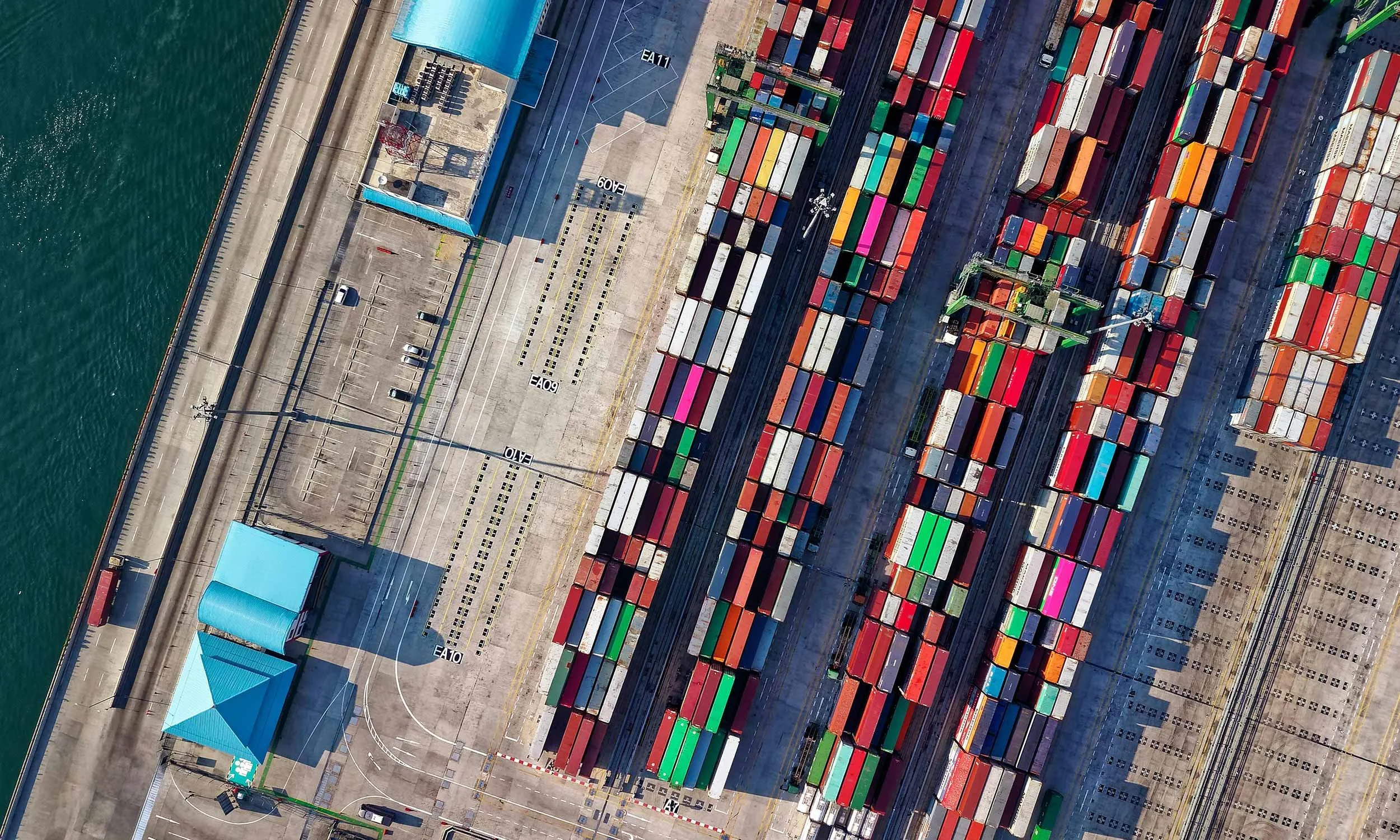 Aerial view of a dock, with colorful shipping containers lined up on railway tracks next to a waterfront port area with parking spaces and a blue-roofed building.