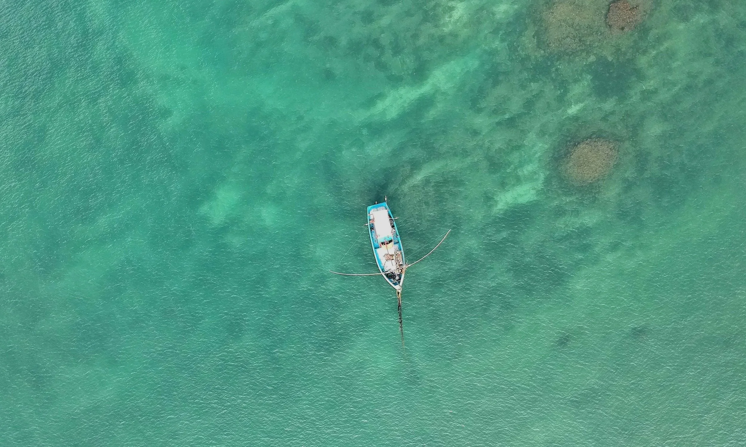 An aerial view of a boat anchored in clear, turquoise water with visible patches of coral beneath the surface.