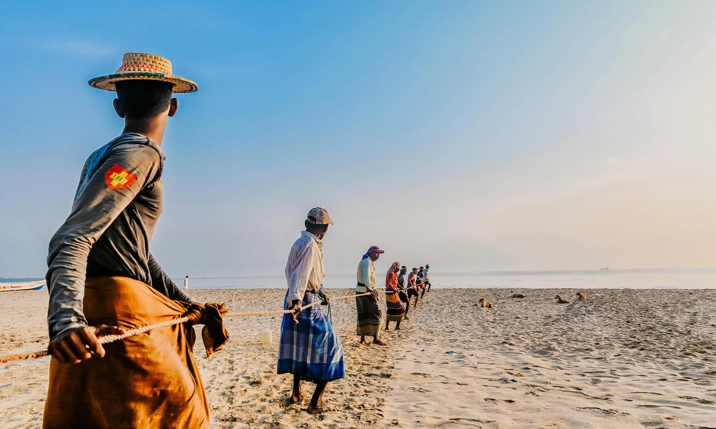 A group of men and boys hauling in a beach seine fishing net, with a calm sea and distant boats in the background.