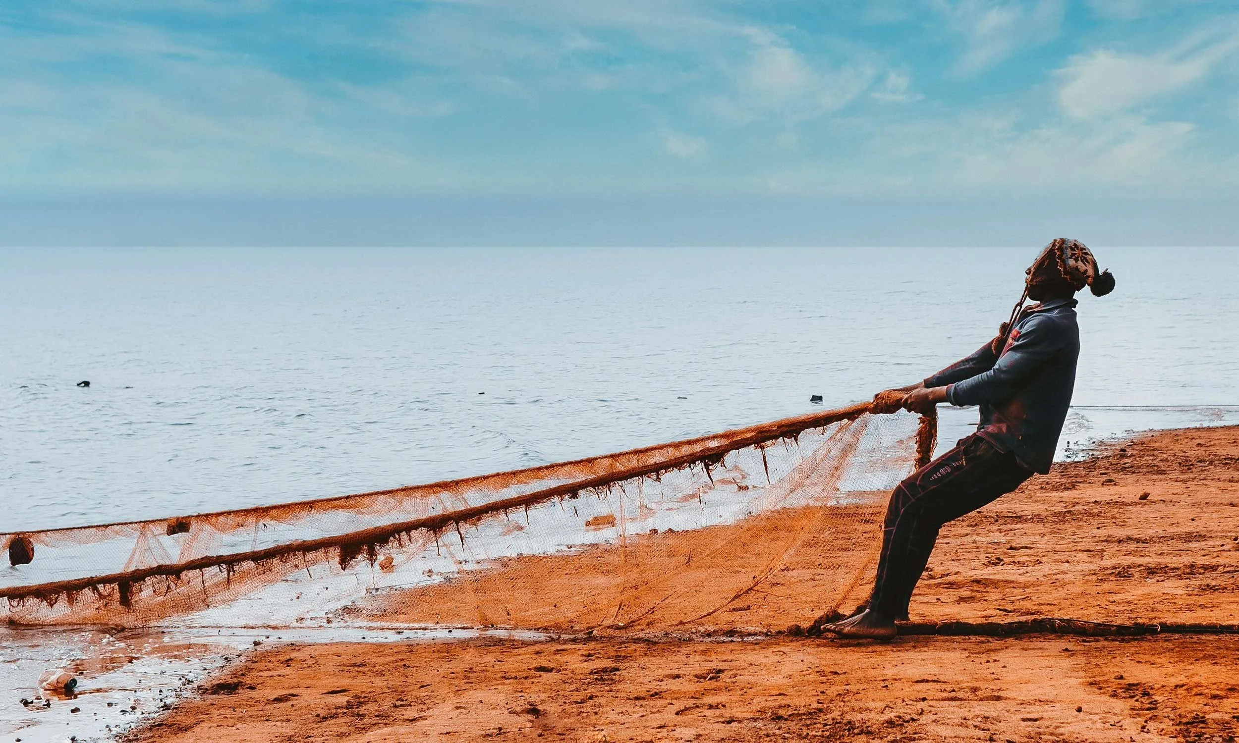 Fisherman hauling a fishing net on beach with ocean and sky in background.