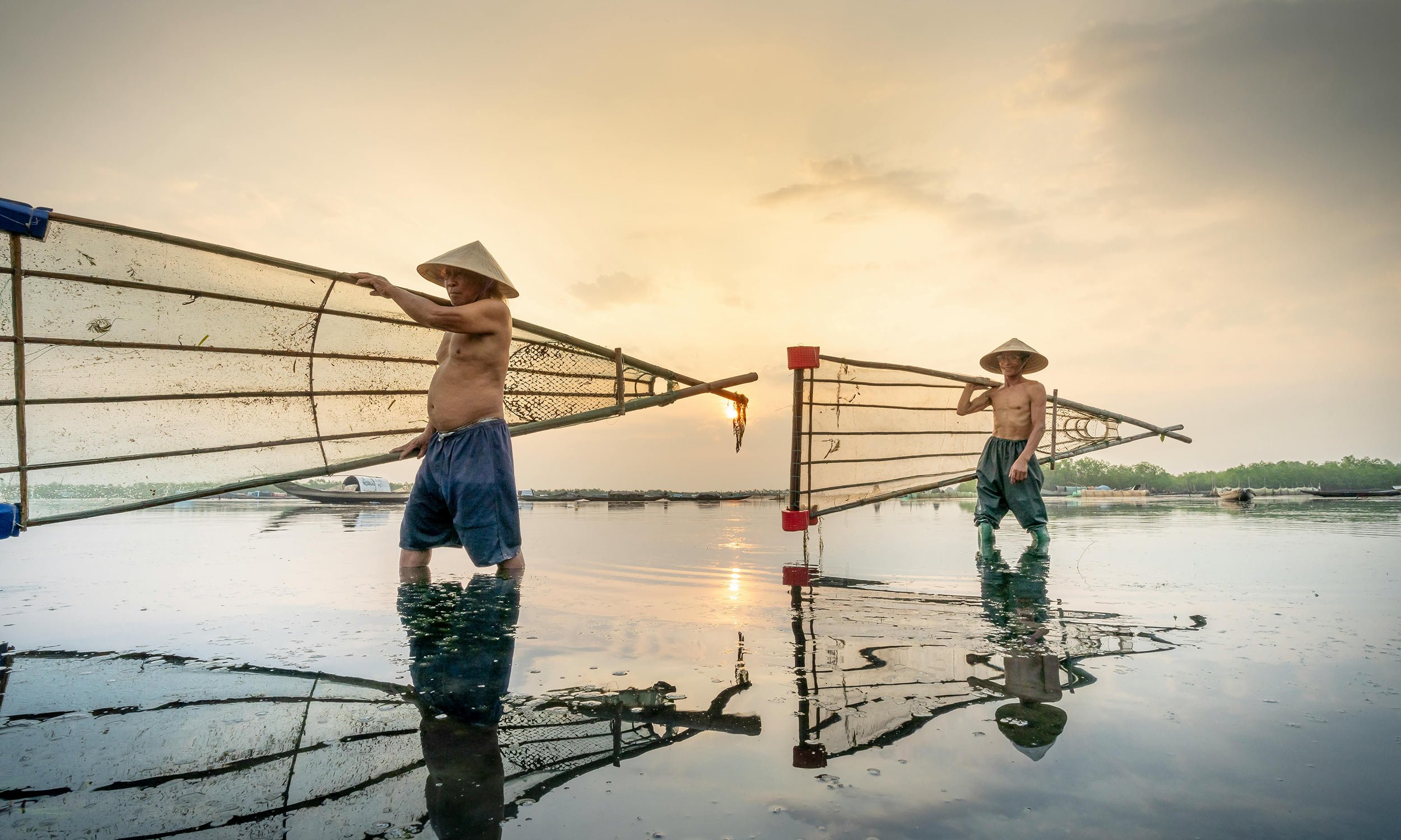 Two shirtless fishermen wearing traditional conical hats wading in shallow water and carrying long fishing nets during sunset.