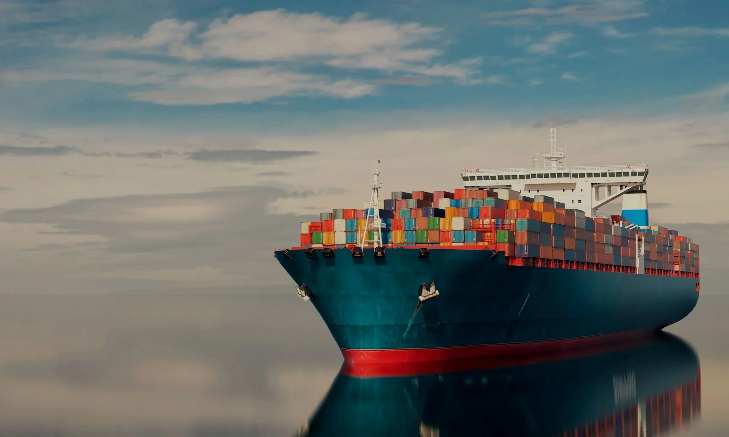 A large cargo ship with colorful containers sailing in calm waters under a partly cloudy sky.