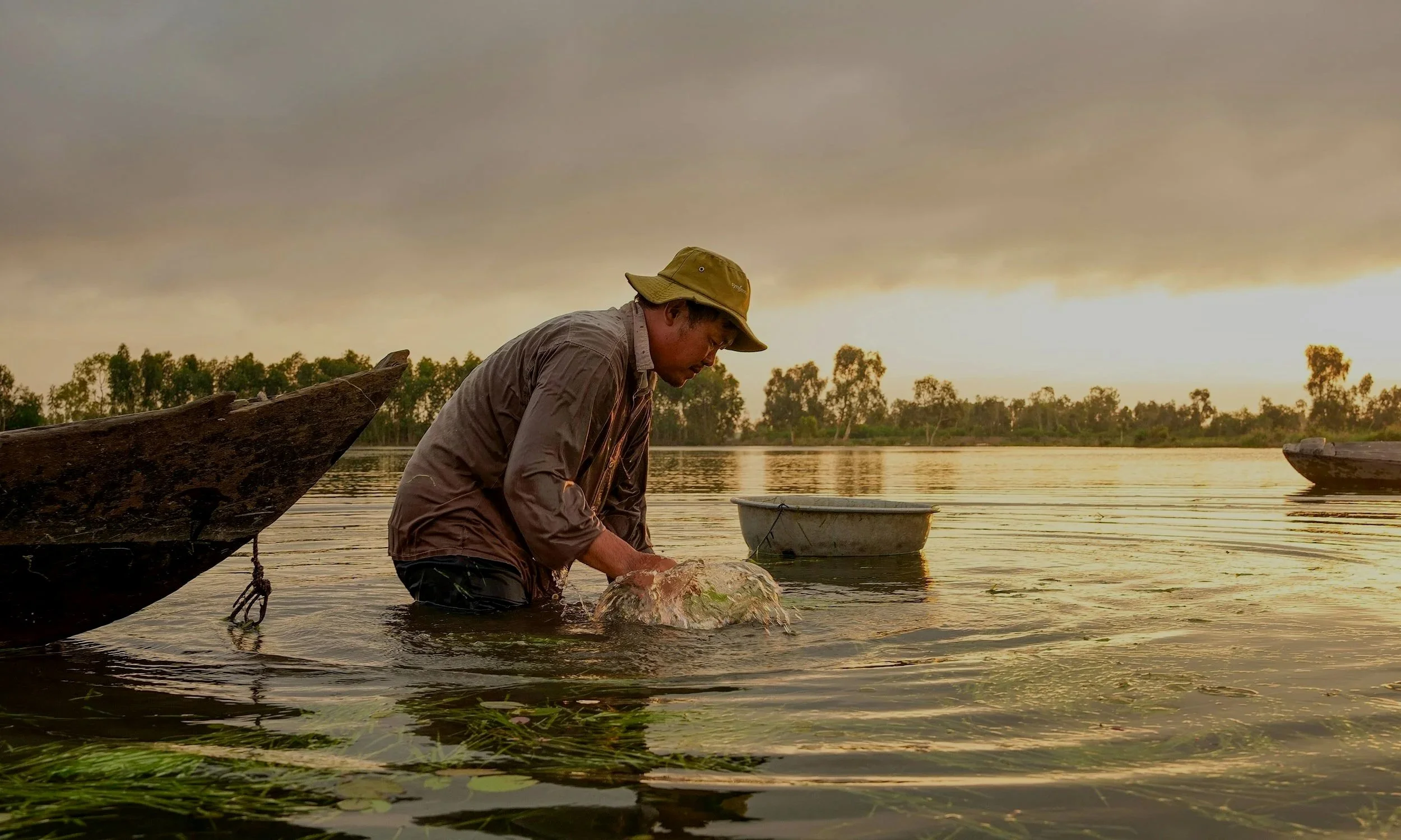 A man in a brown jacket and wide-brimmed hat is standing in a body of water, holding a large fish. Two boats are nearby in the water, with a backdrop of trees and a cloudy sunset sky.