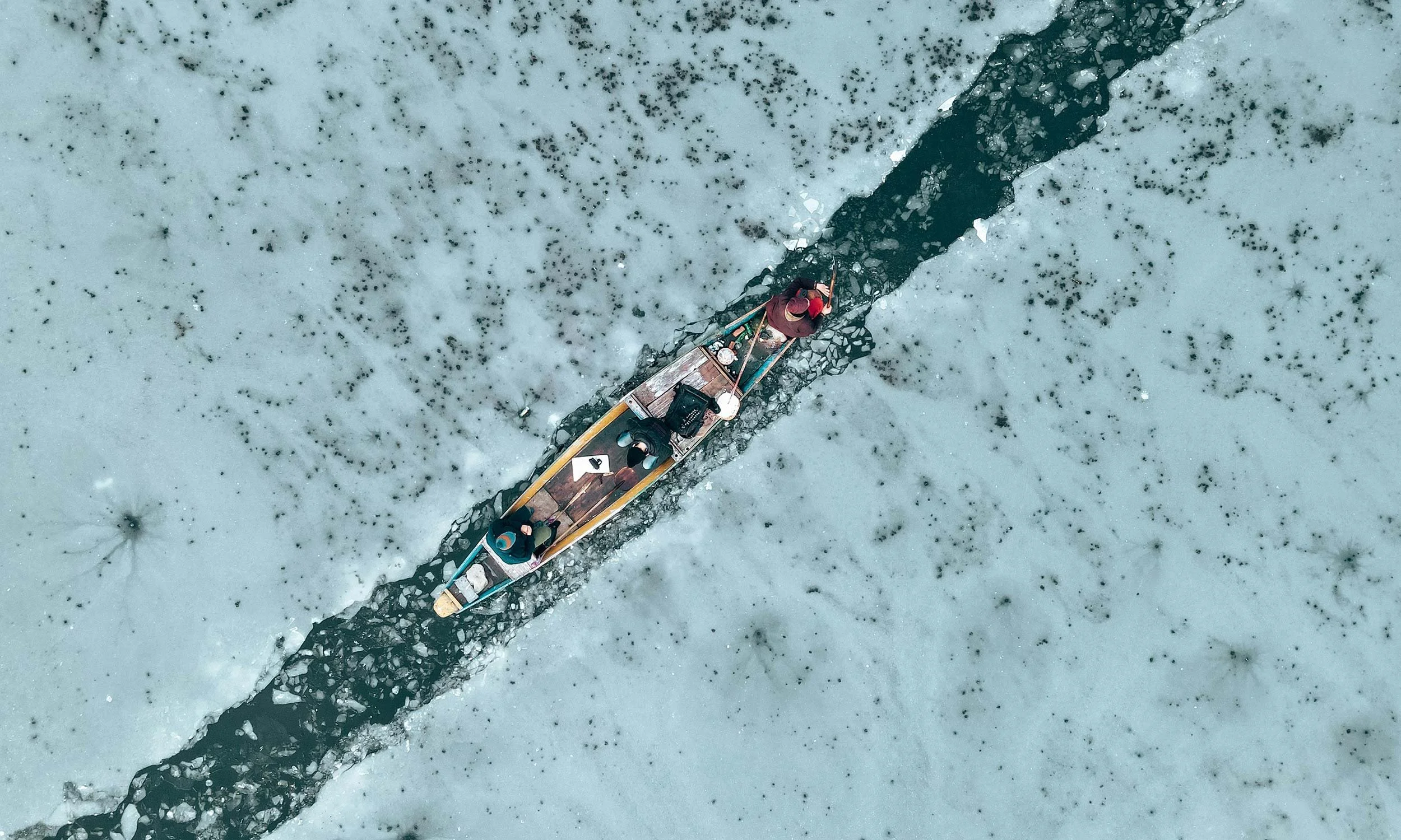 An aerial view of a boat with three people on a narrow black ice trail through a frozen, snow-covered body of water.
