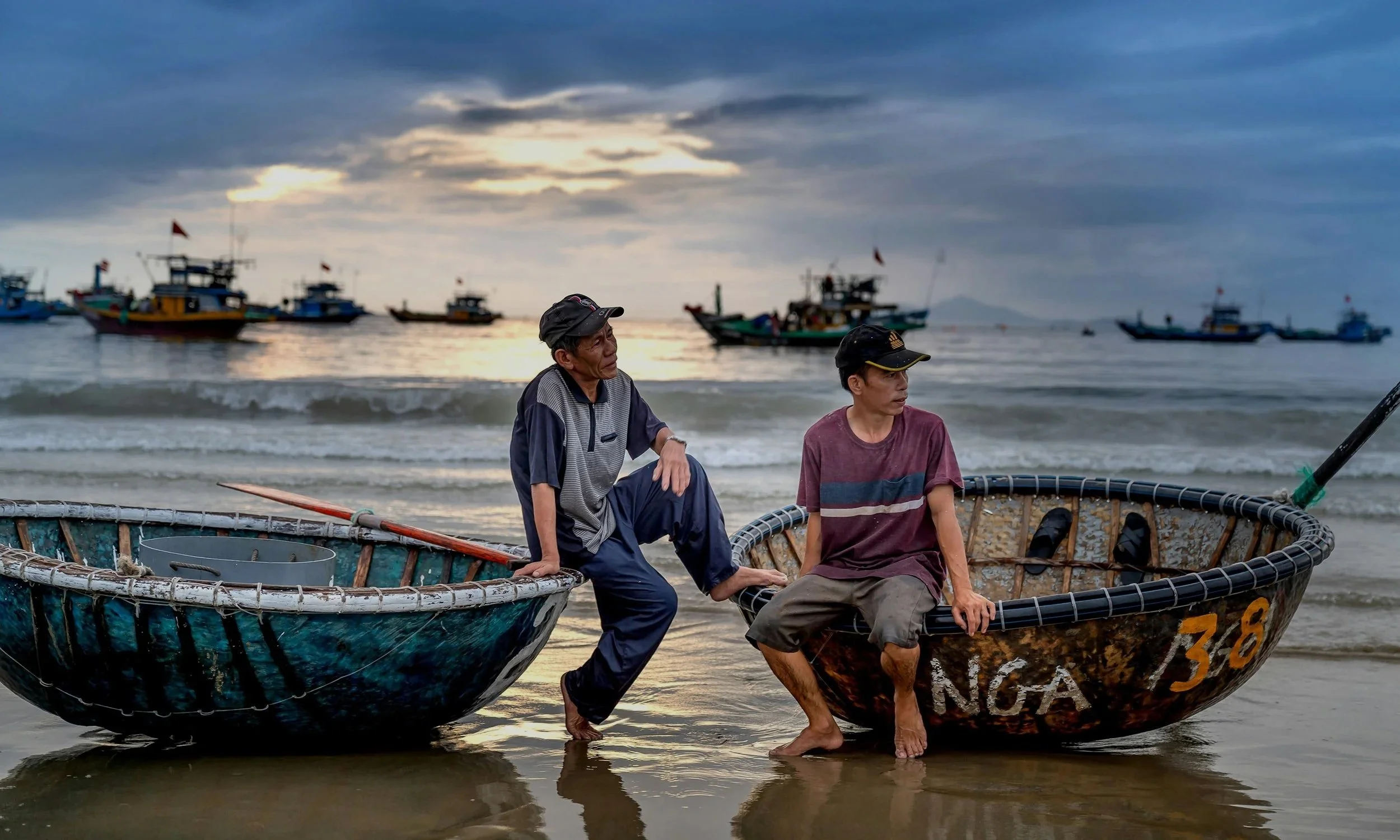 Two fishermen sitting on traditional round basket boats on a beach at sunset, with fishing boats anchored in the background under a cloudy sky.