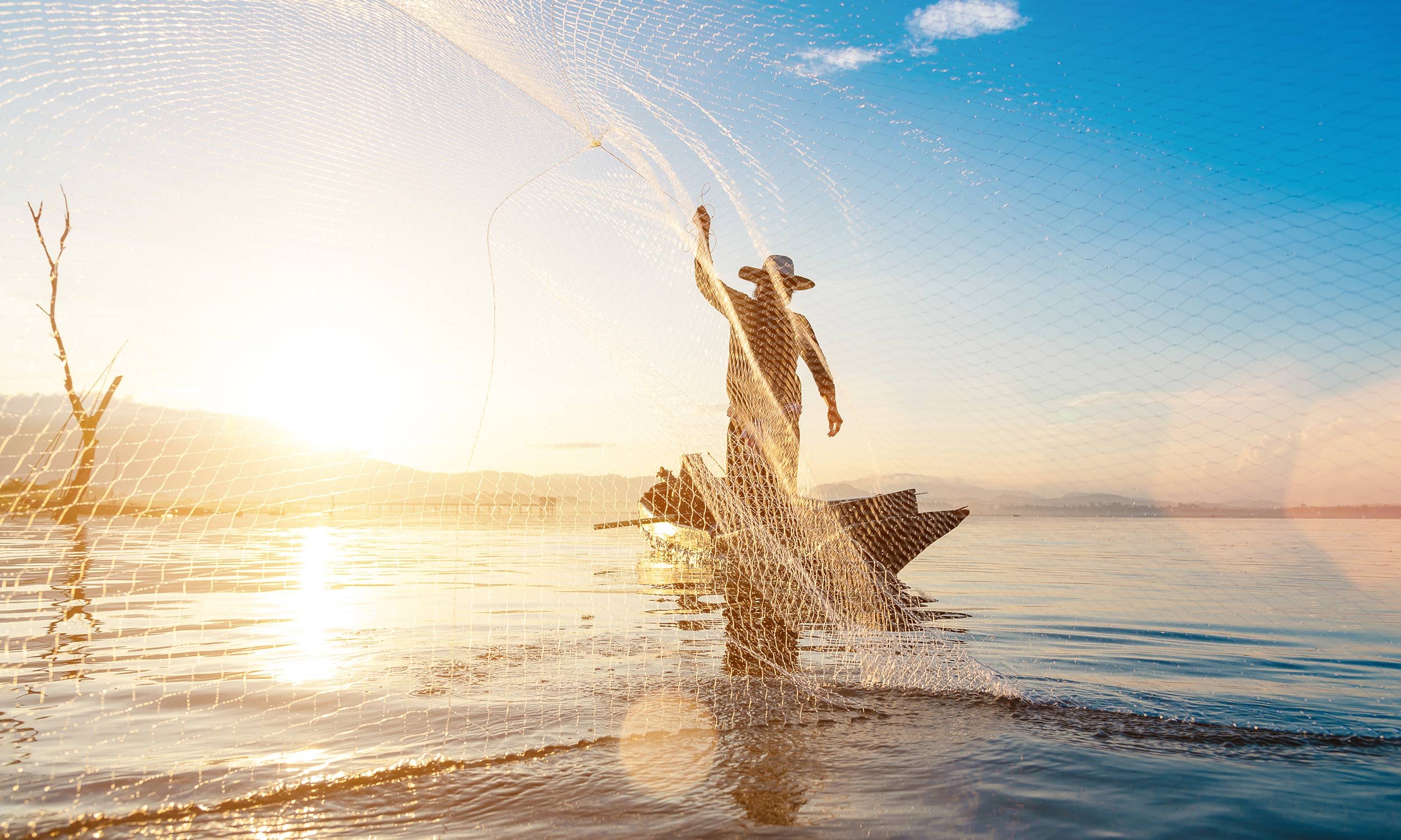Fisherman casting a fishing net into calm waters at sunrise or sunset, with a clear sky and distant mountains.