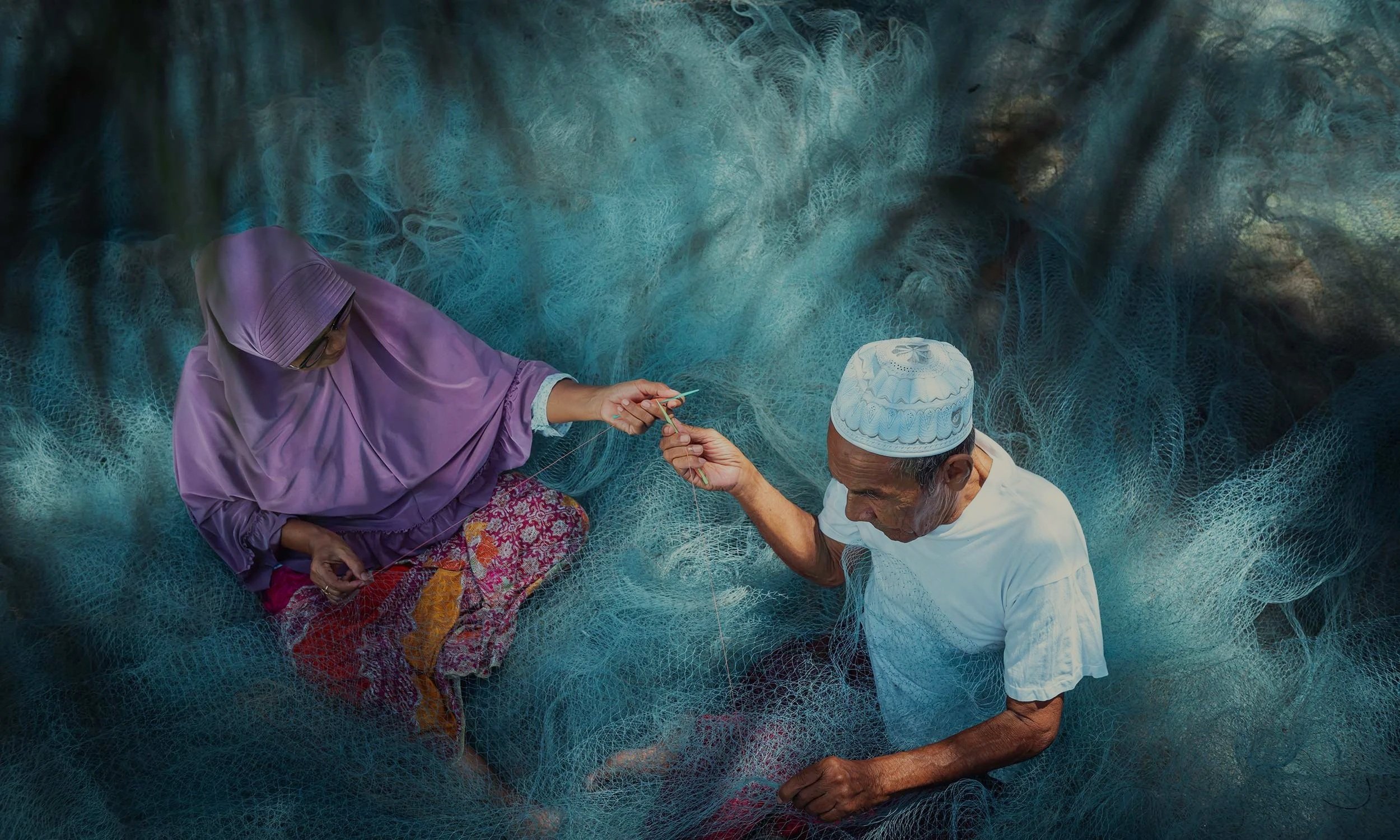 Two elderly people, a man and a woman, are repairing fishing nets. The woman is wearing a purple hijab, while the man is wearing a white shirt.
