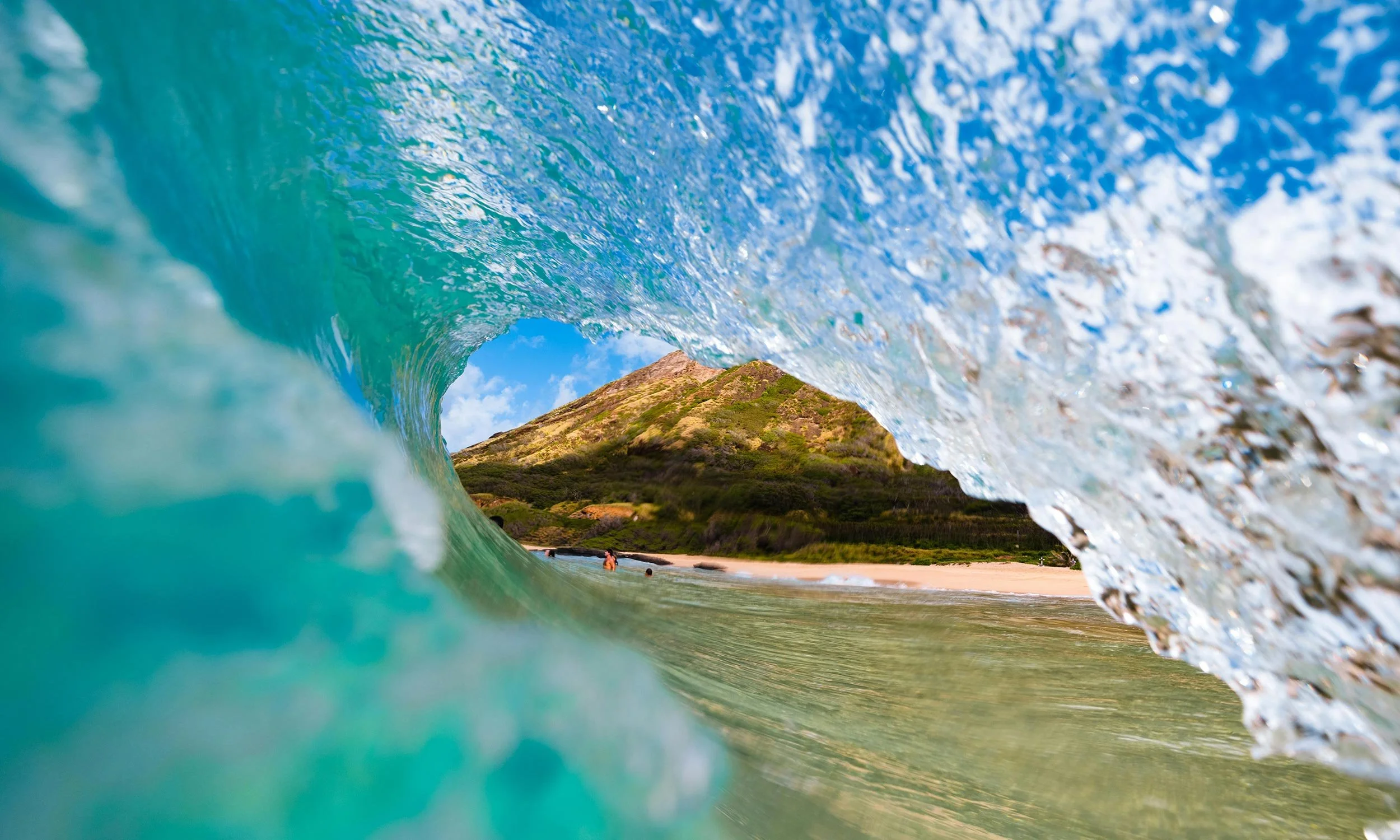 Photo of a wave crashing on a tropical beach with a mountain in the background and some people in the water.