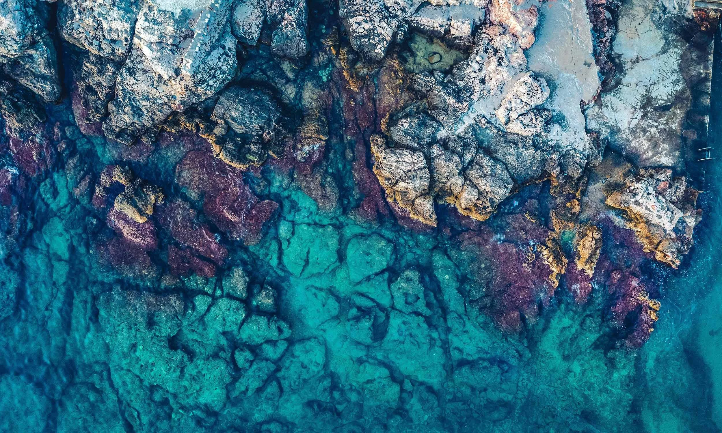 Aerial view of a rocky coastline with clear blue water and some rocks with vibrant purple, gold, and white hues.