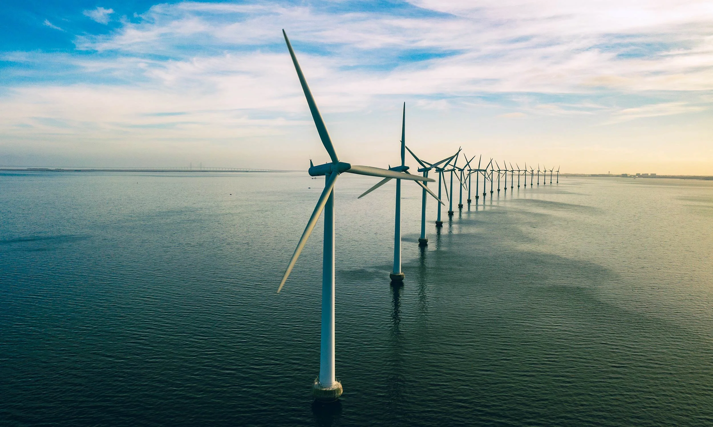 A row of offshore wind turbines in the water extending into the horizon under a partly cloudy sky.
