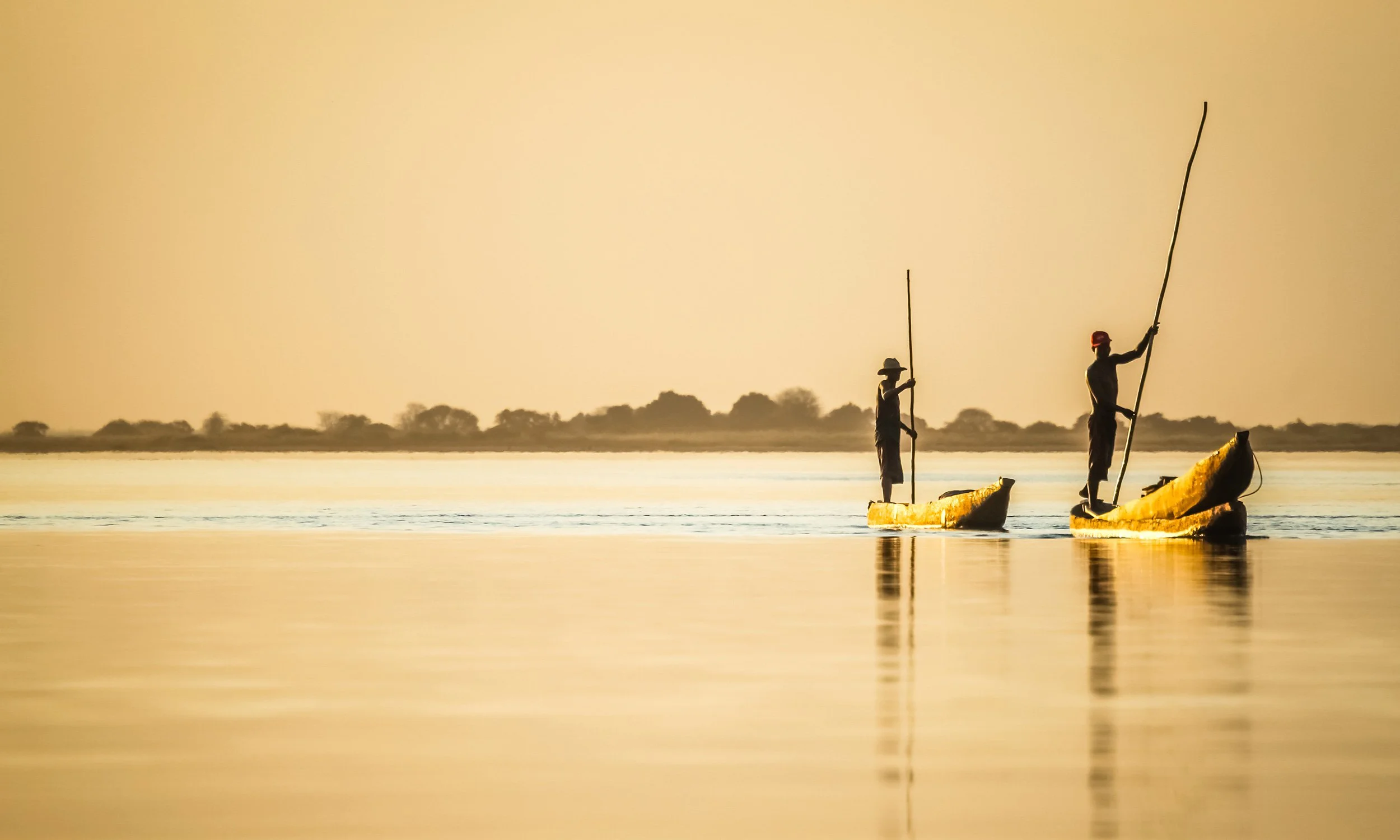 Two people stand on yellow boats in calm water, holding long poles, with a flat landscape in the background during a golden sunset.