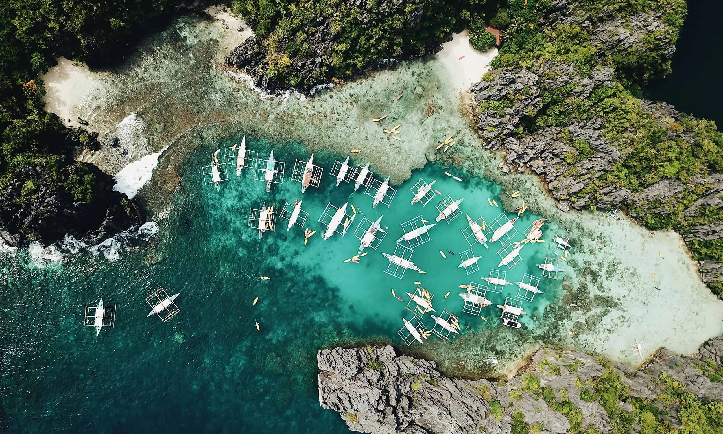 Aerial view of a cove with turquoise water, surrounded by lush green cliffs. Several traditional wooden boats are floating in the water near the sandy beach.