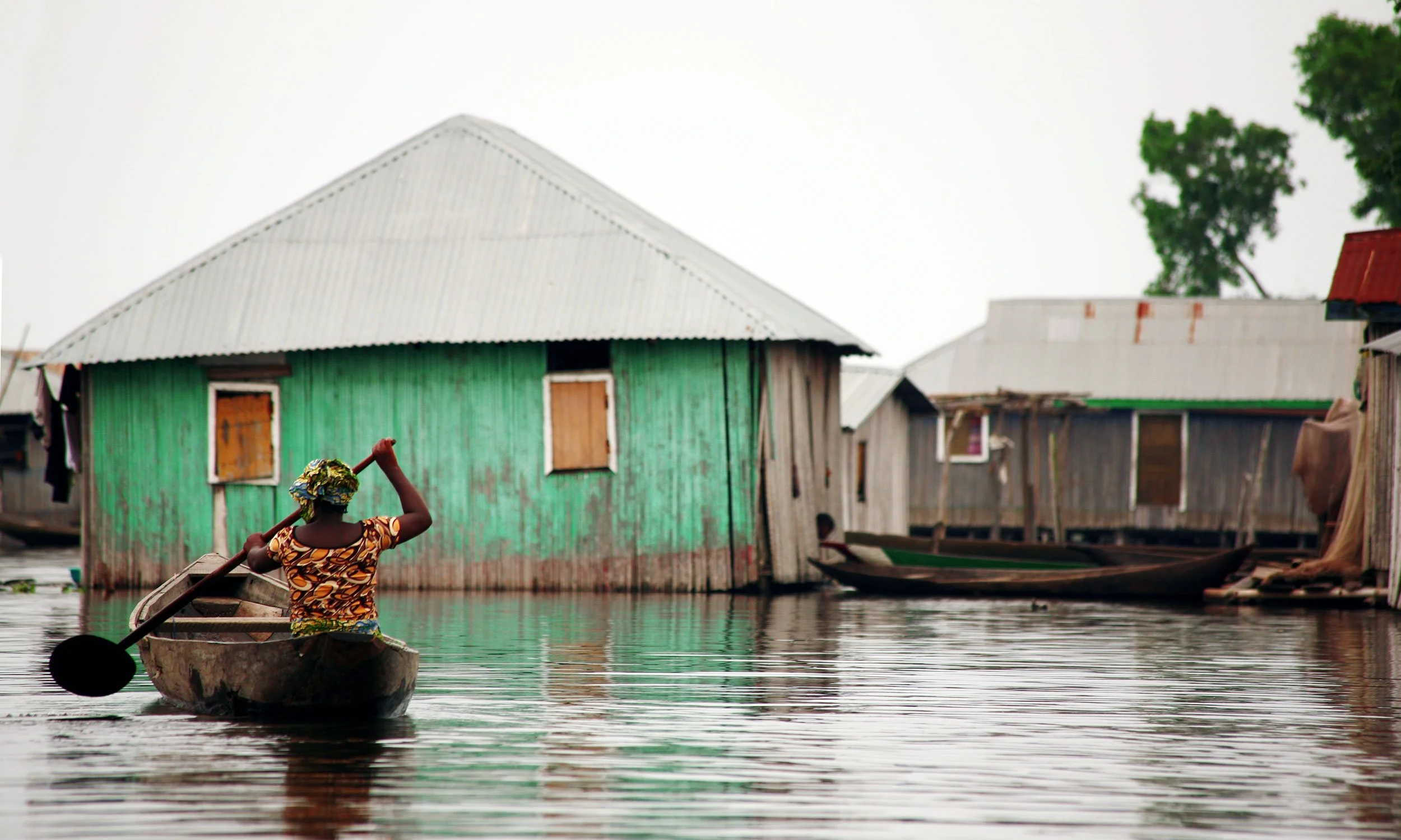 A person paddles a canoe as waters rise around houses on stilts in the background.