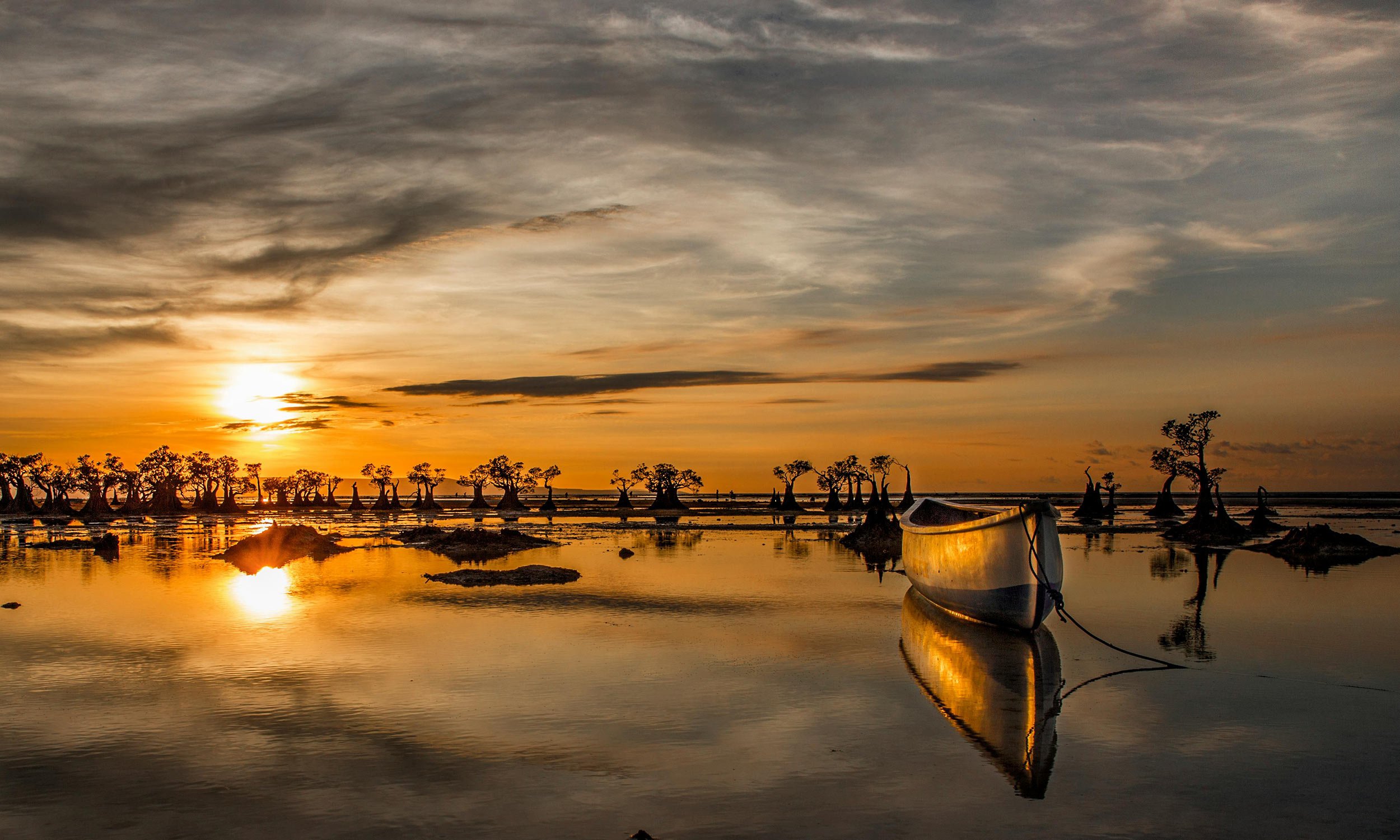 Sunset over calm water with a traditional fishing canoe and silhouettes of mangrove trees in the distance.