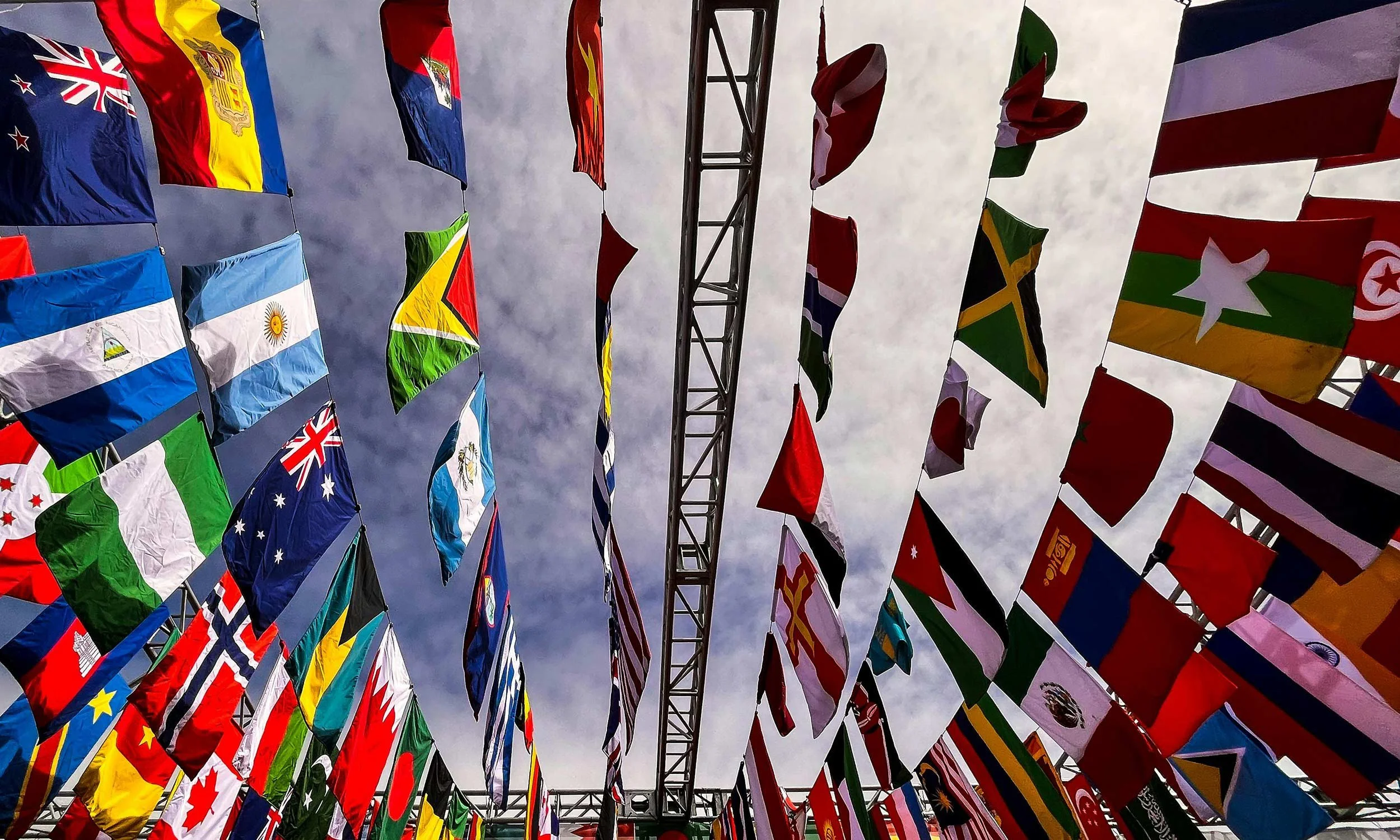 Multiple country flags hanging on strings against a cloudy sky, including flags of Australia, Argentina, Guyana, Bangladesh, Tanzania, Namibia, Zimbabwe, and others.