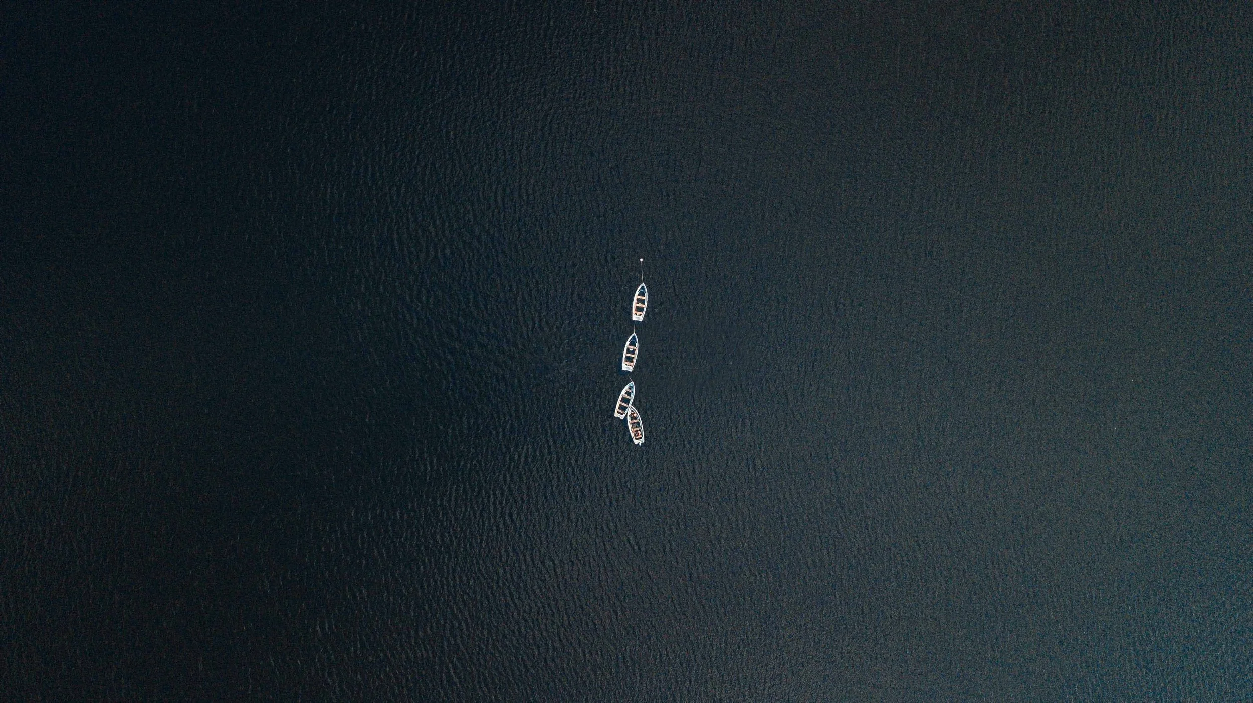 Five boats are anchored in a line on calm dark water.