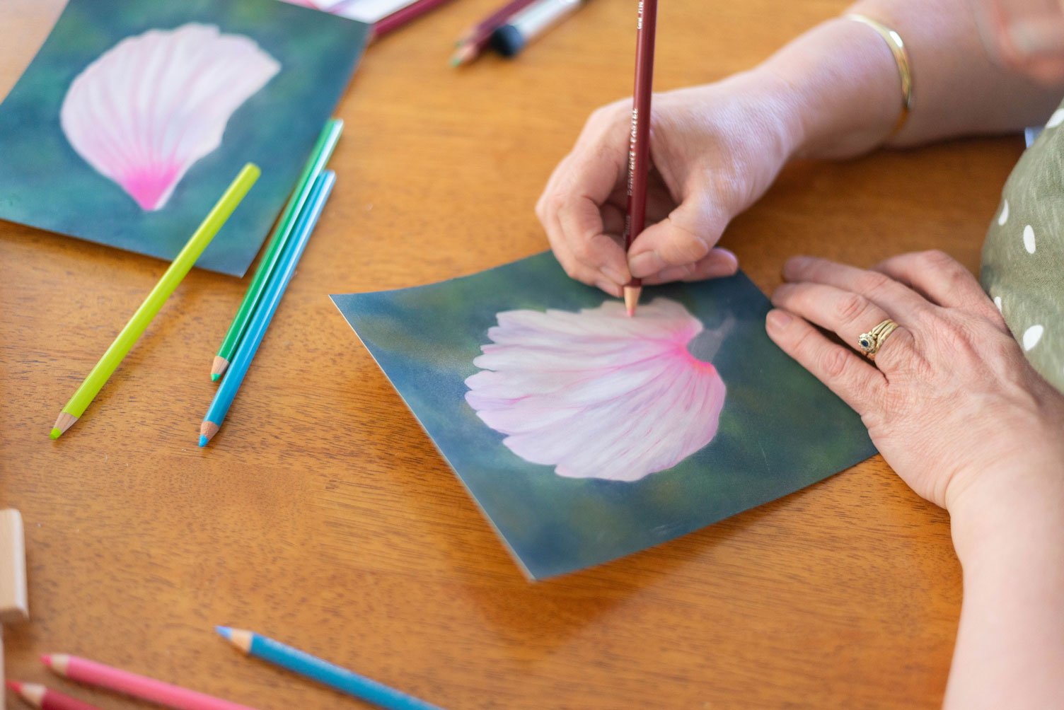 A person is using colored pencils to create a pastel drawing of a pink and white flower on a dark background, with more colored pencils and a reference photo of the flower nearby on a wooden table.
