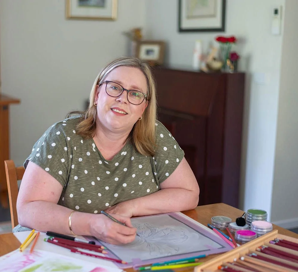 A woman with glasses and shoulder-length blonde hair sitting at a table, coloring a drawing with colored pencils.