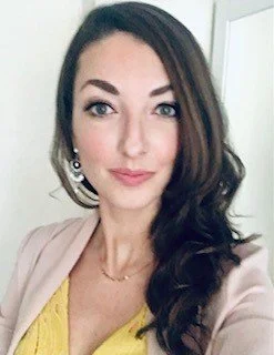 A woman with long, wavy dark hair, wearing makeup, earrings, a beige blazer, and a yellow top, posing indoors.