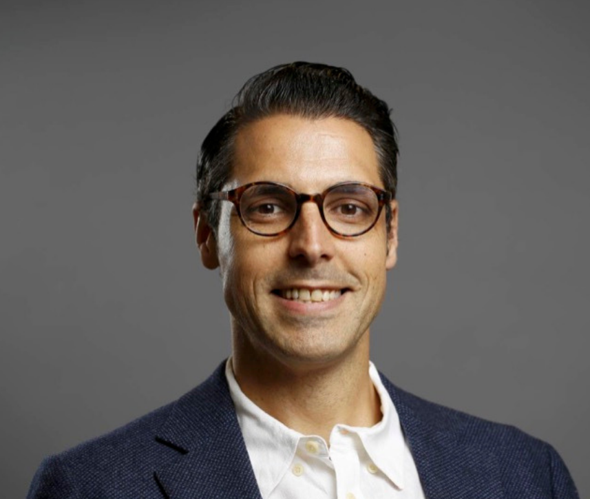A man with dark hair, wearing glasses, a white shirt, and a blue blazer, smiling against a gray background.