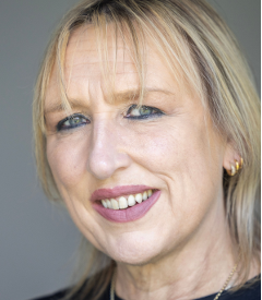 Close-up portrait of a blonde woman with short hair, smiling against a neutral background.