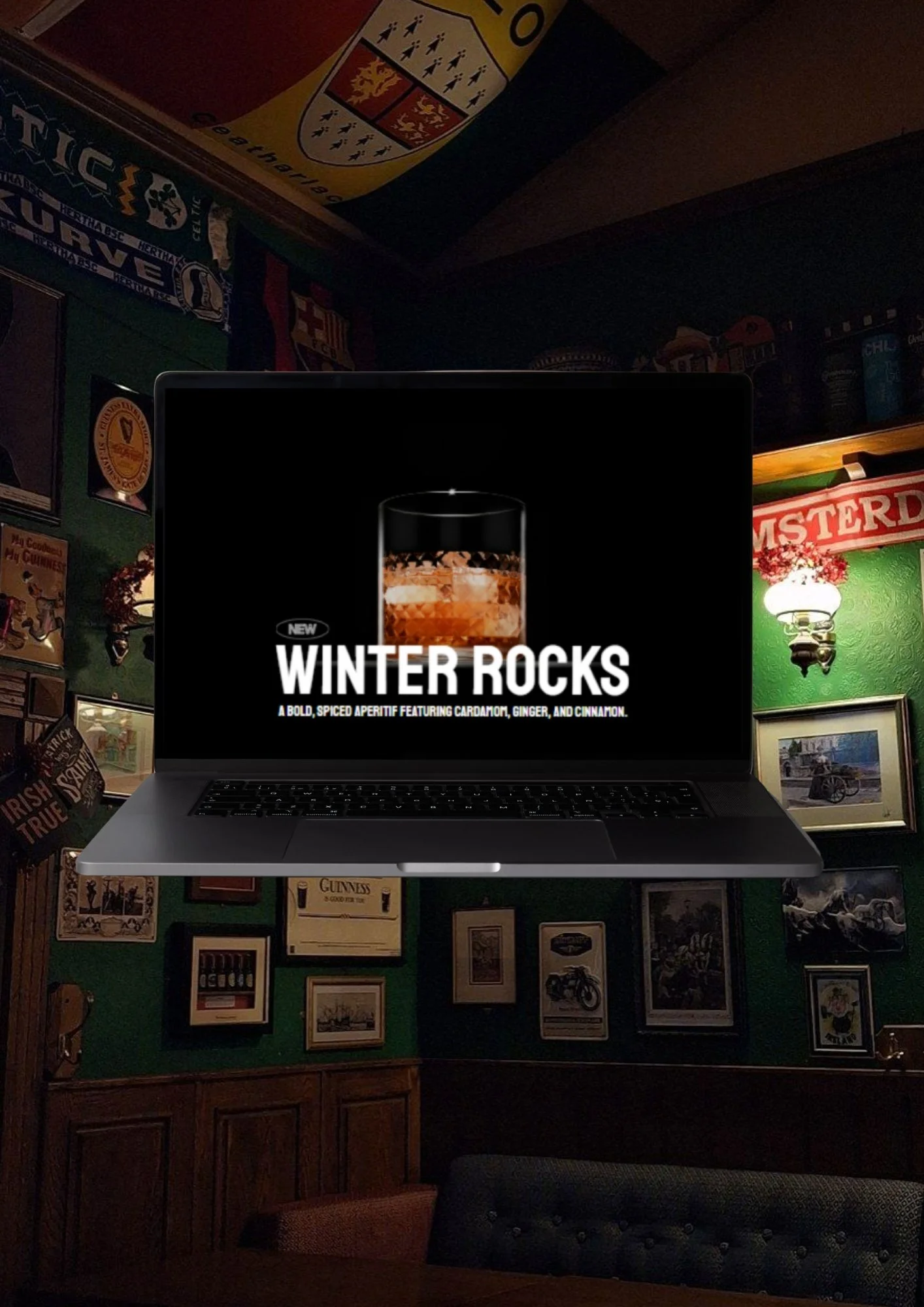 Laptop displaying an advertisement for Winter Rocks, a bold, spiced aperitif with cardamom, ginger, and cinnamon, placed in a pub with Irish and British-themed decorations on the walls.