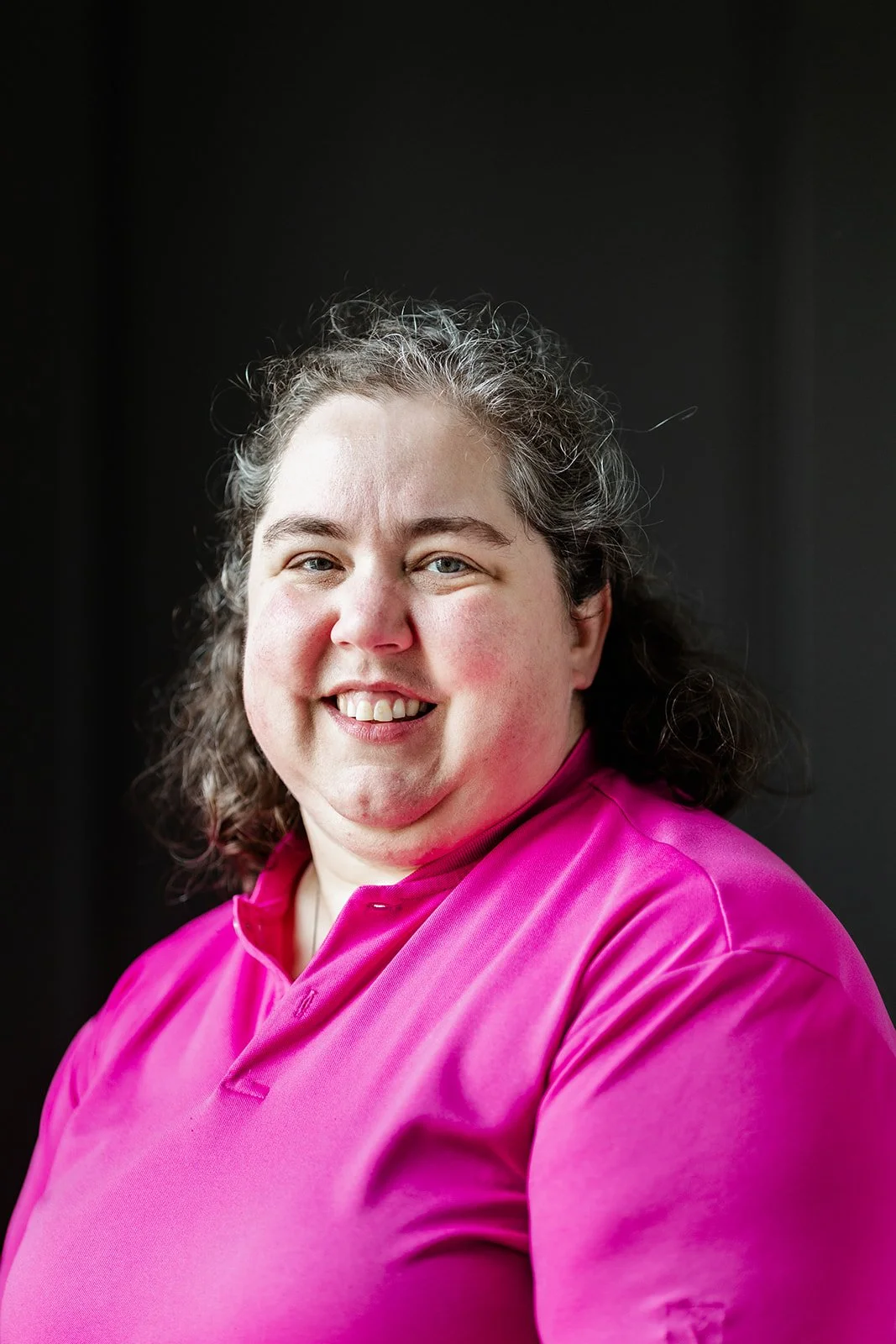 A woman with curly brown hair, wearing a black shirt and red jacket, smiling at the camera against a plain white background.