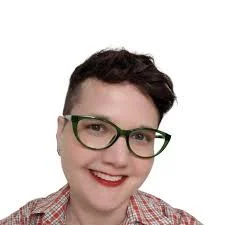 Close-up of a person with short dark hair, wearing green glasses, red lipstick, a small hoop earring, and a plaid shirt, smiling against a plain white background.
