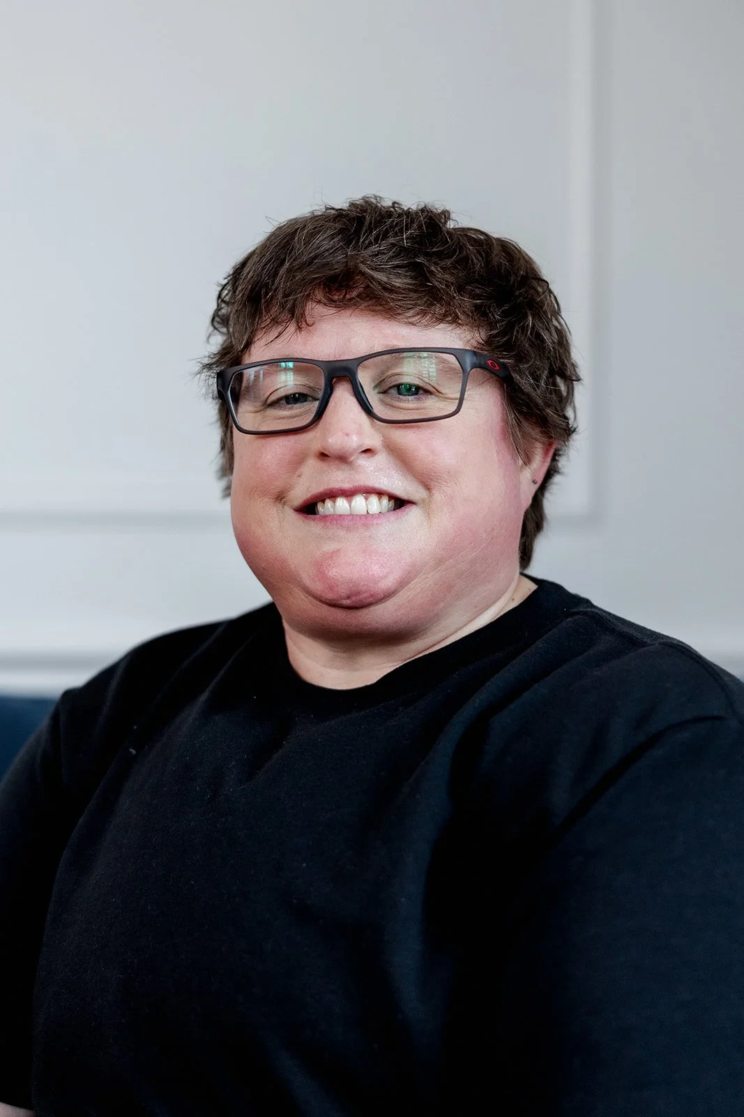 Close-up of a woman with tousled light brown hair, blue eyes, wearing a black shirt, smiling gently, against a plain white background.