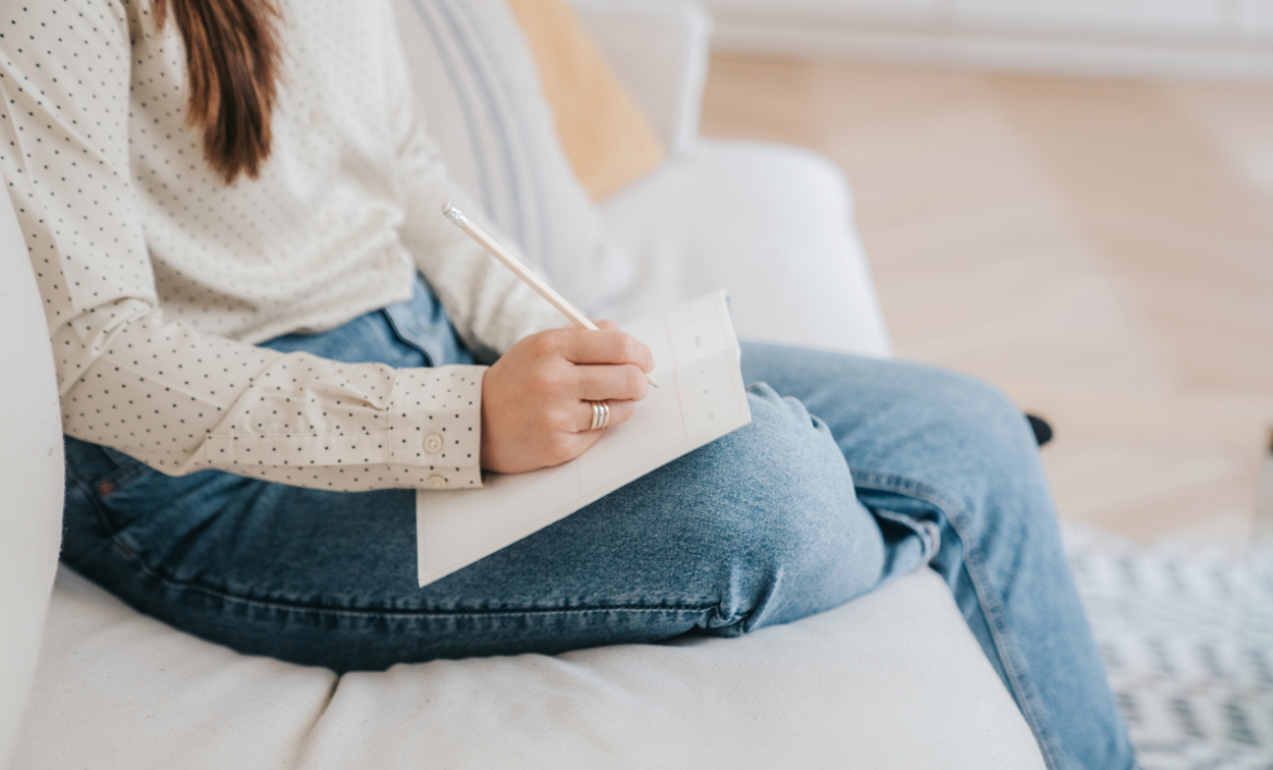 A woman sits on a sofa wearing jeans and a polka dot shirt. She is writing in a notepad.