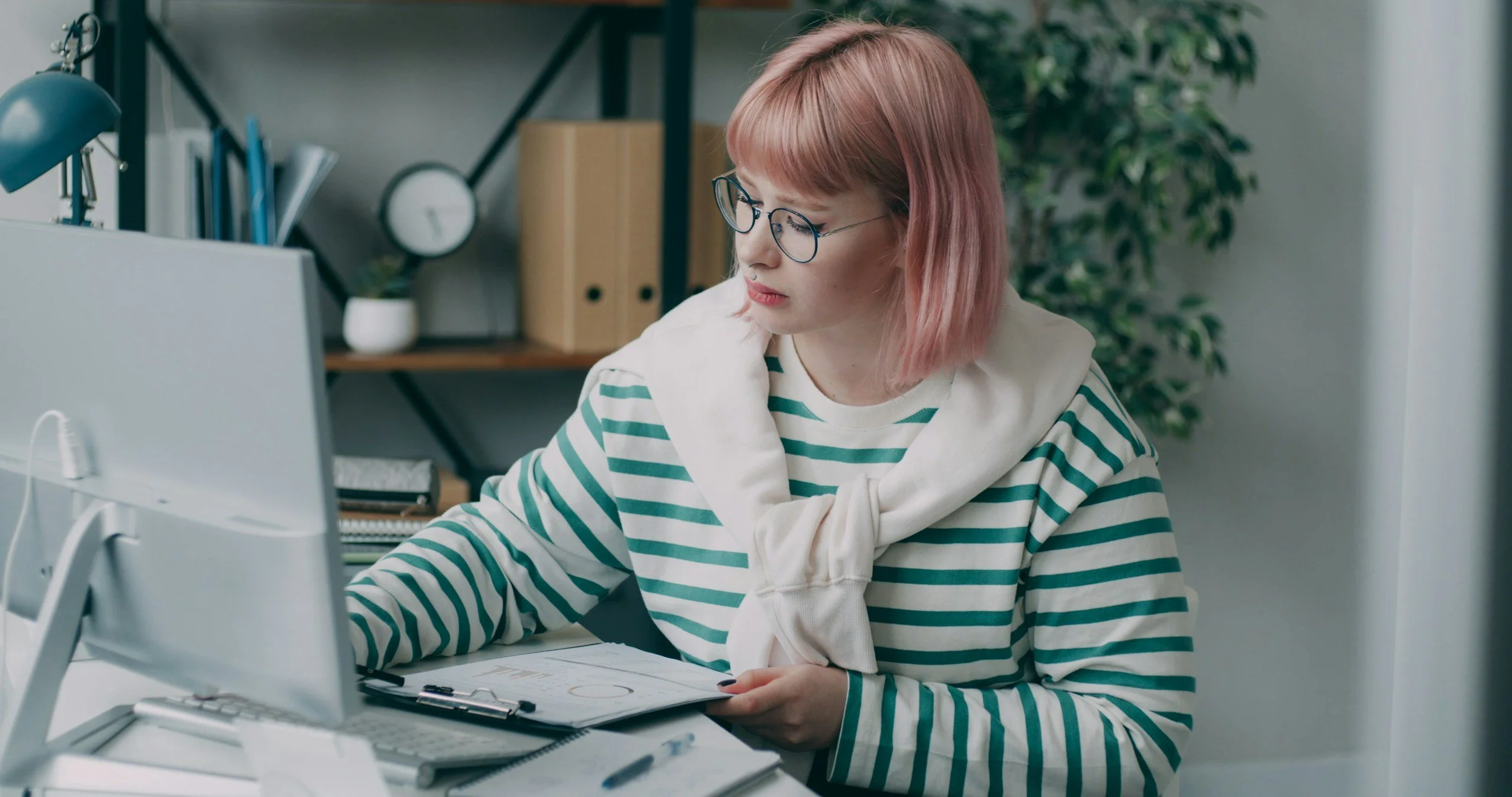 A woman with pink hair wearing a green and white striped top sits in front of a computer monitor working. Psychodynamic therapist in Surrey helping women stop repeating relationship patterns online across the UK.