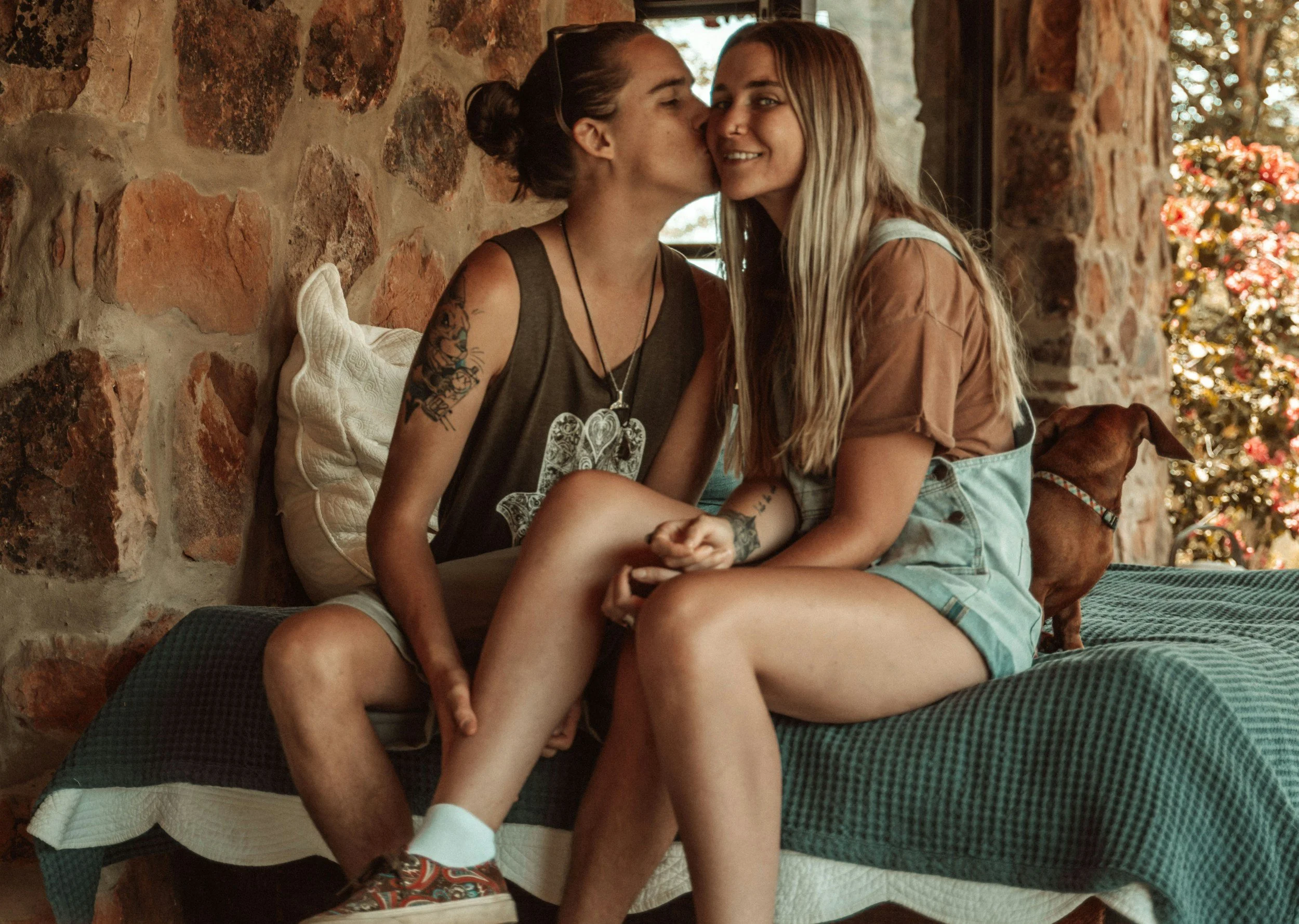 Two women sitting close together beside some brickwork, one kissing the other on the cheek while the recipient of the kiss smiles at the camera.