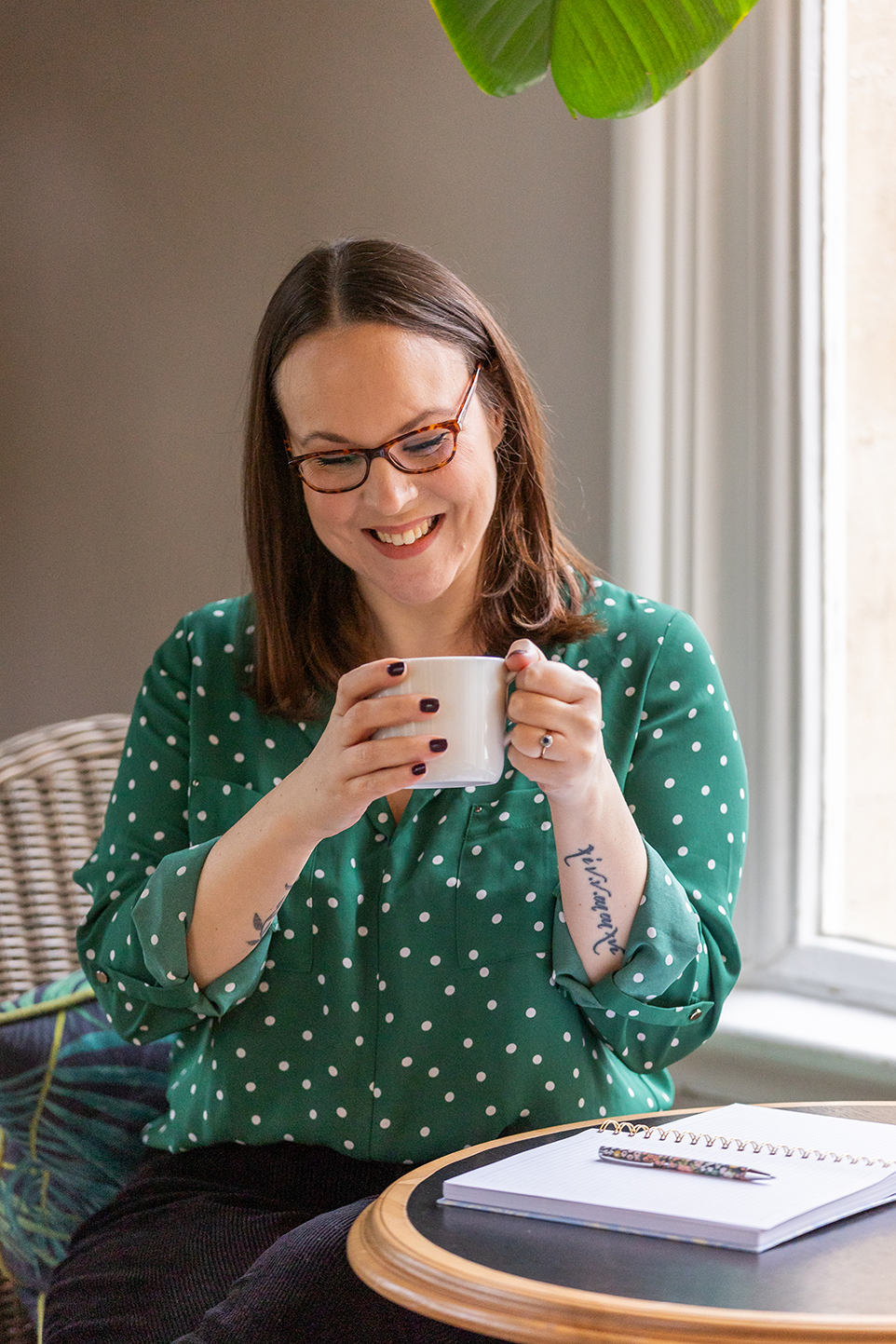 A woman with dark hair wearing a green top with white polka dots is holding a white mug and smiling while looking down