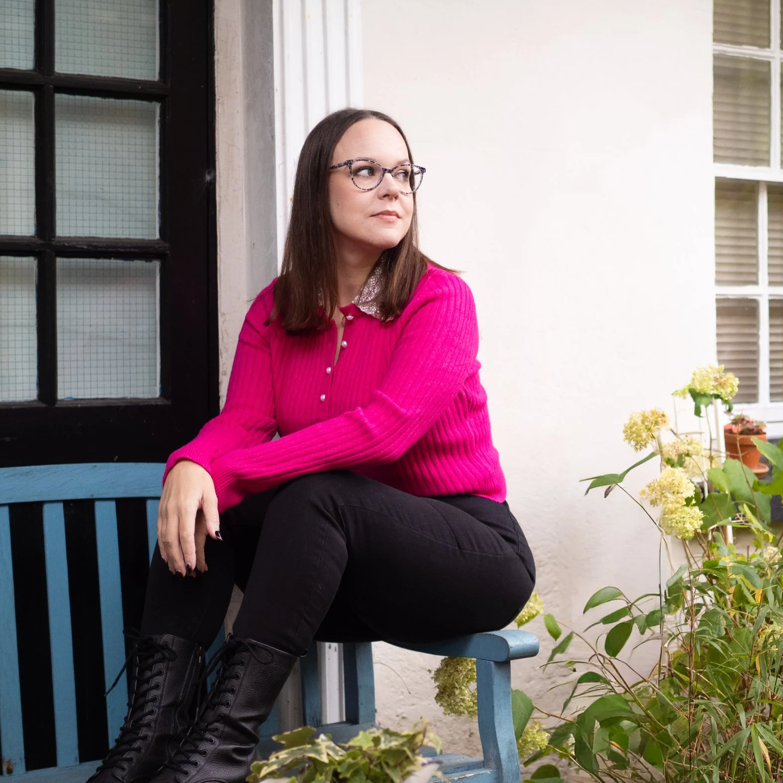 A woman with brunette hair wearing a pink jumper and black jeans sits on the arm of a turquoise bench next to a plant. She is looking in to the distance with a slight smile on her face