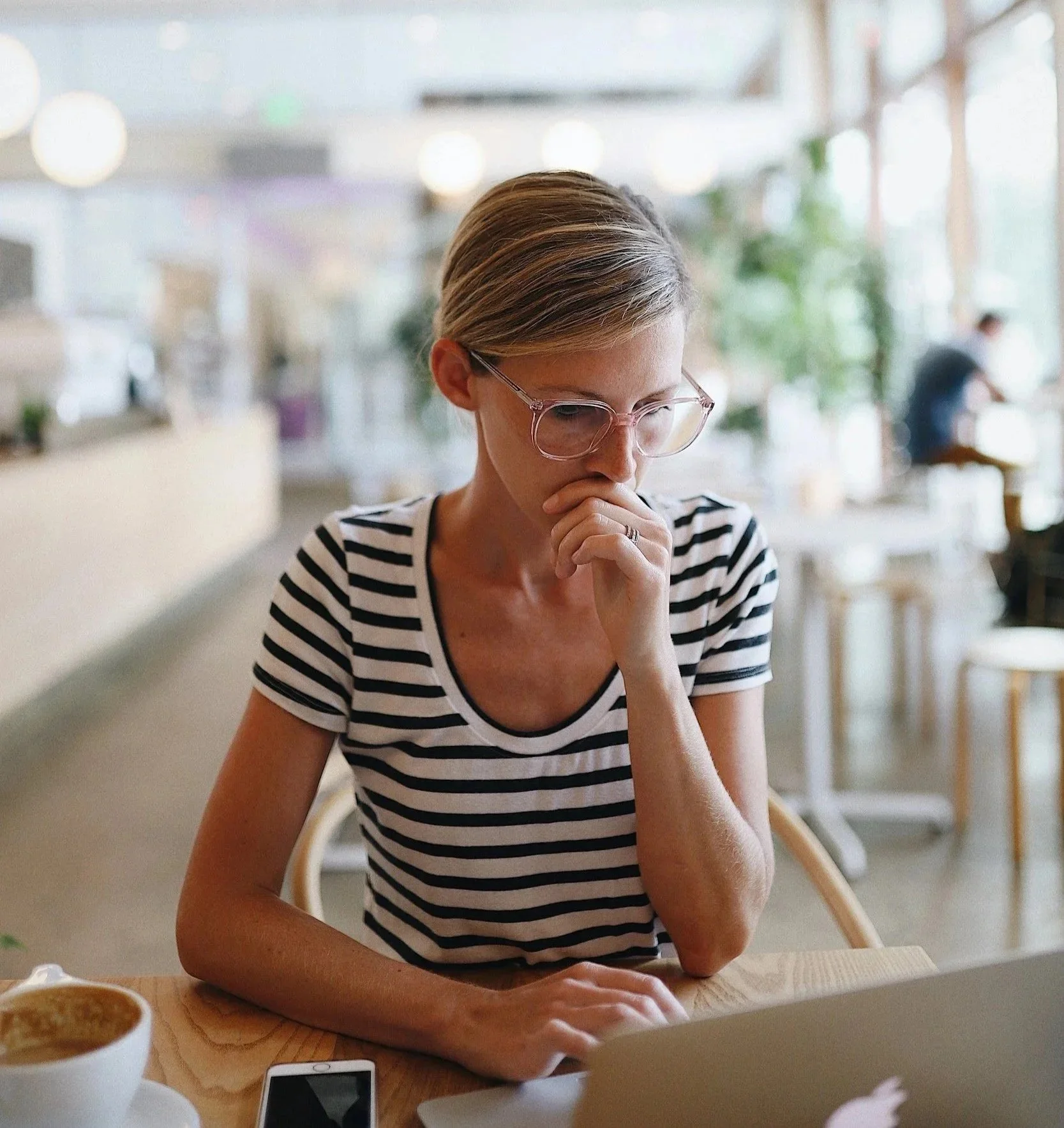 A blond woman wearing glasses and a black and white striped t-shirt sits in front of an open laptop with a worried look on her face