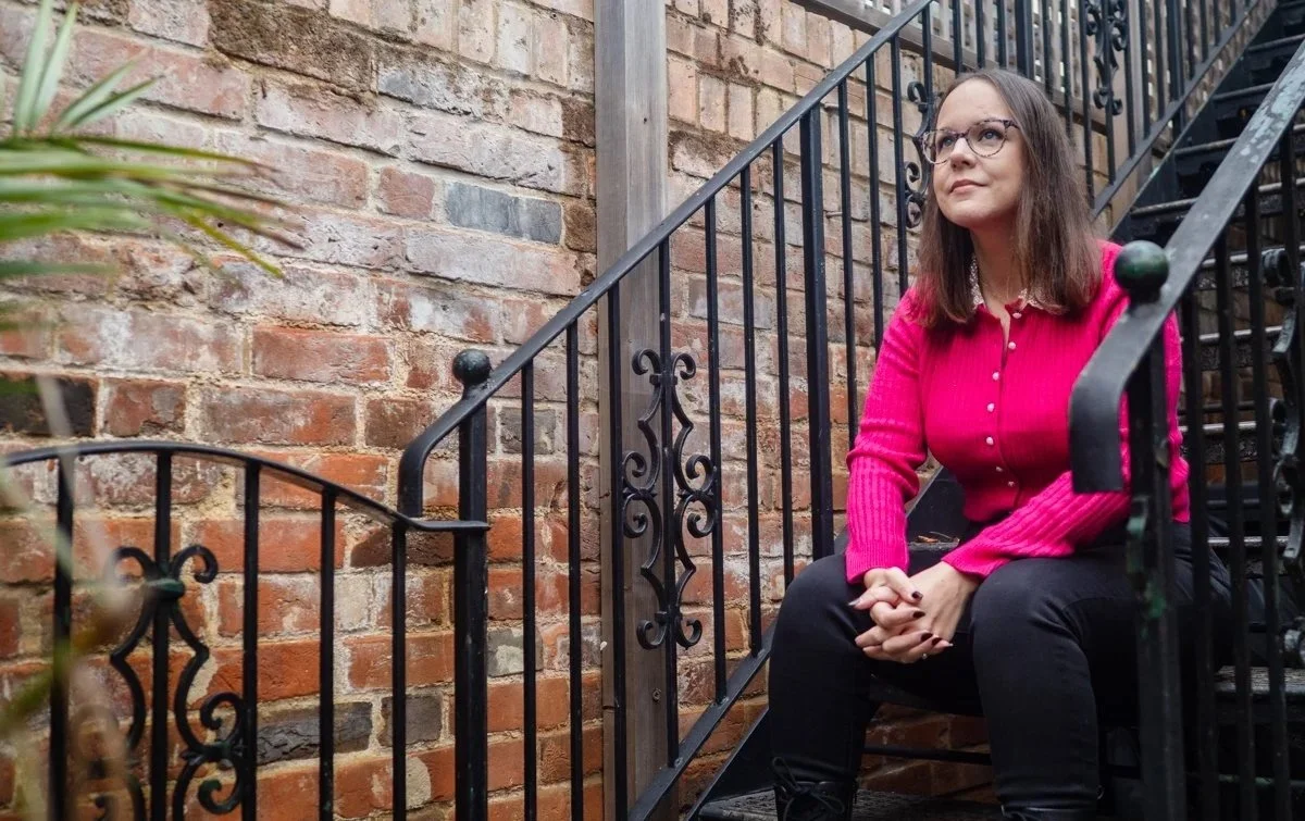 Aimee Bishop, a woman with glasses sitting on an outdoor metal staircase with a brick wall background, wearing a pink sweater and black pants. Therapy with Aimee. Counselling in Bisley Woking Surrey.