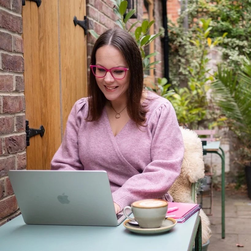 Aimee Bishop, a woman with dark hair, pink glasses, and a pink sweater is sitting outside at a table with a laptop, a cup of coffee, and a notebook, smiling while working. Therapy with Aimee. Counselling in Bisley Woking Surrey.