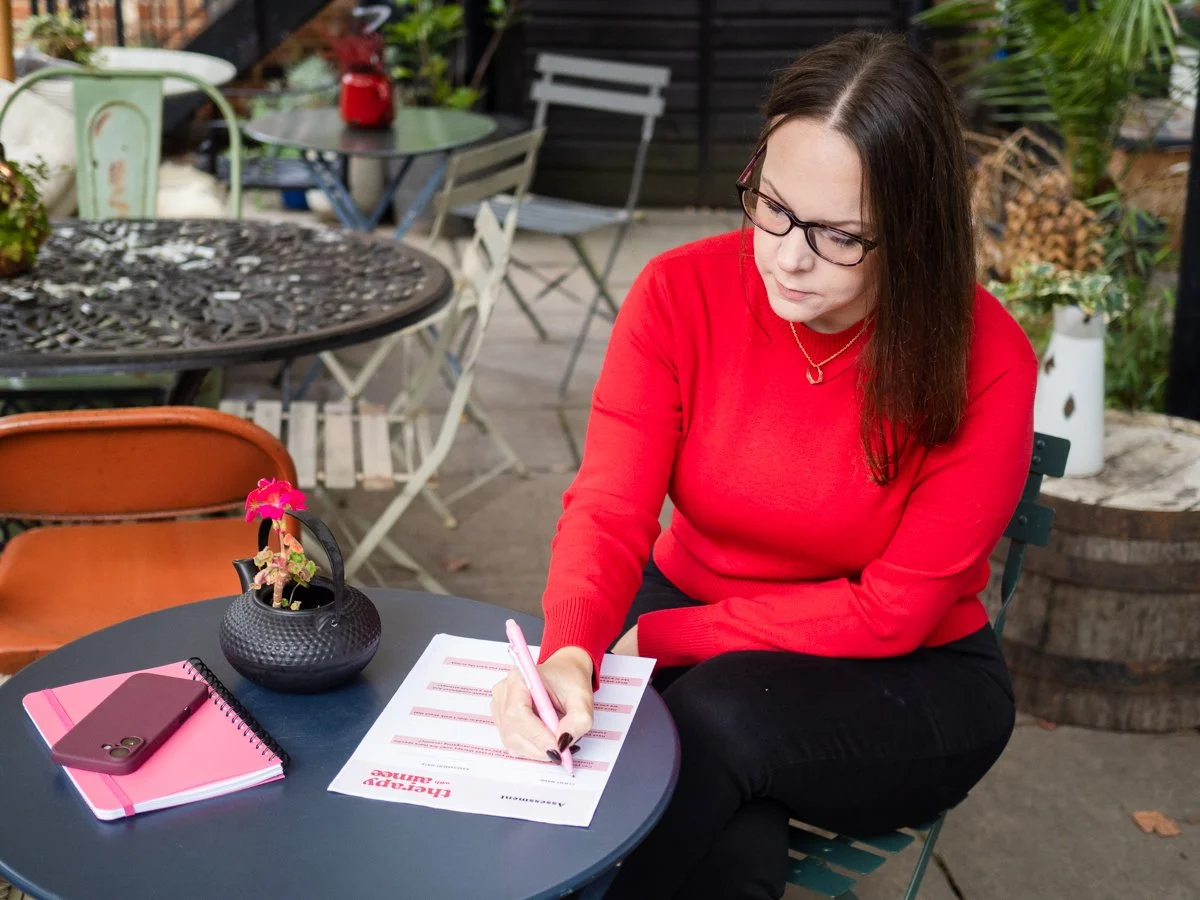 Aimee Bishop, a brunette woman in red sweater sitting at outdoor cafe table, is writing notes with a pink pen, surrounded by pink notebook, smartphone, and potted plant.