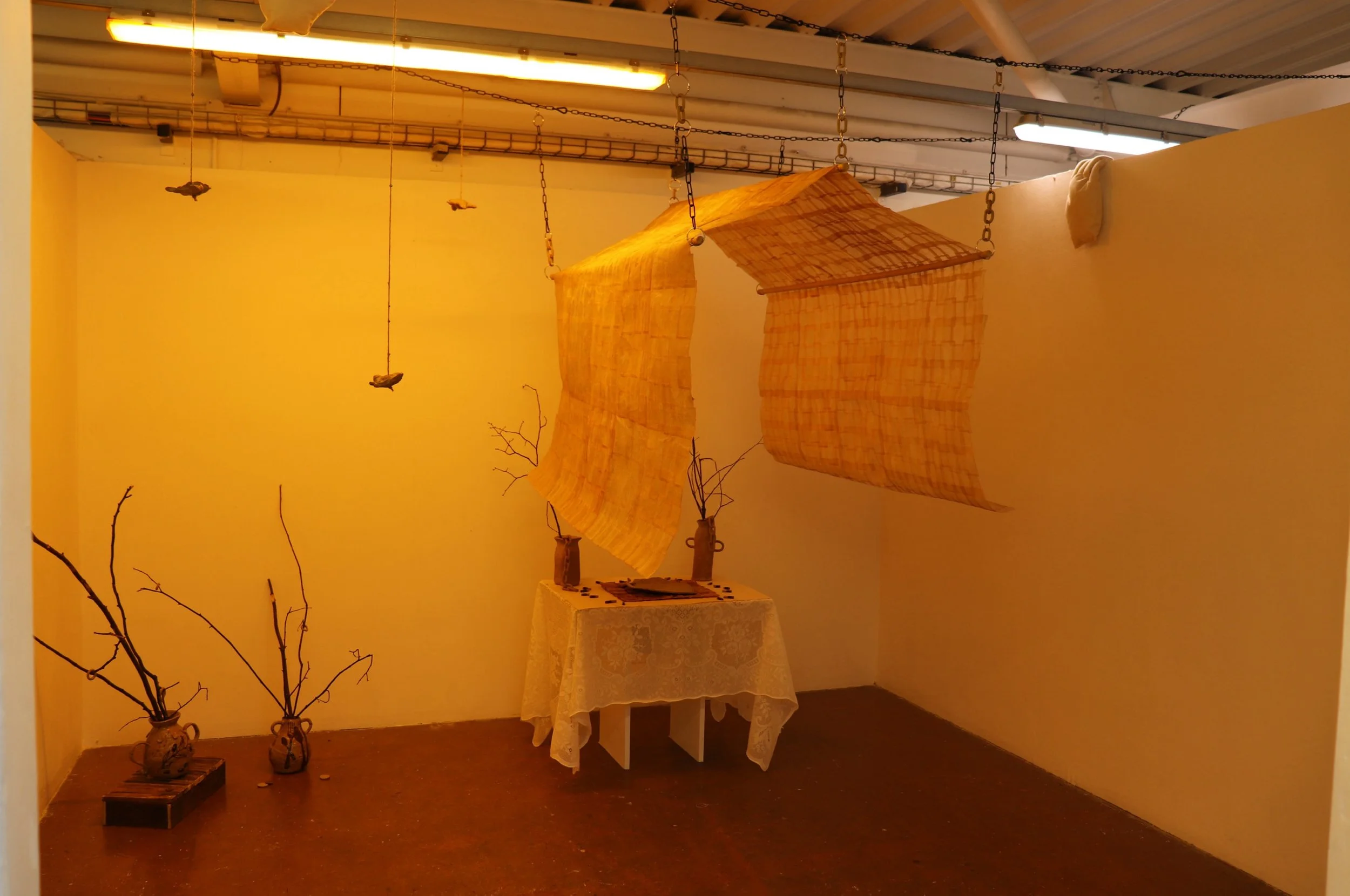 An art installation featuring hanging fabric sheets suspended by chains, with a table covered with lace cloth beneath, and two bare branches in vases on the floor, all set against a warm yellow wall.