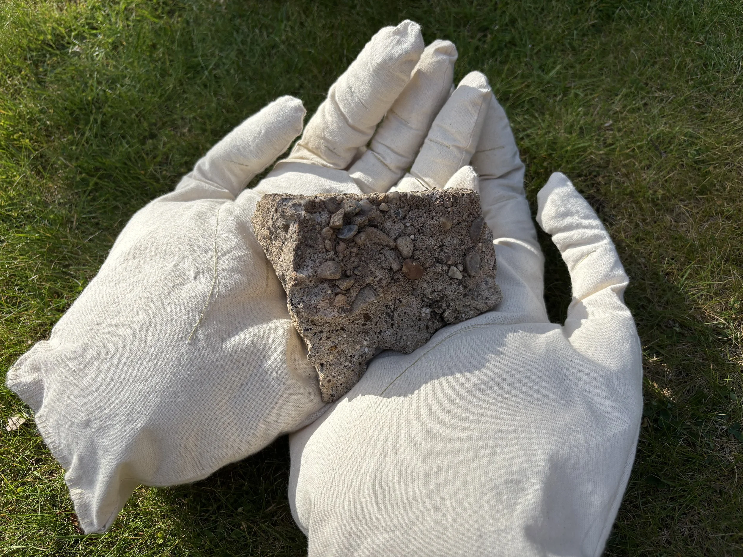 A person wearing white gloves holding a piece of concrete with small rocks embedded in it over green grass.