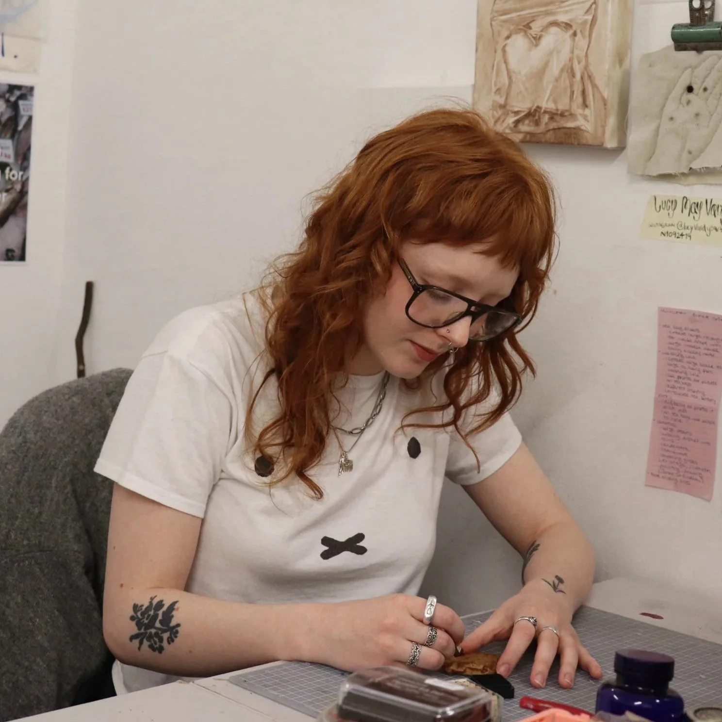 Red-haired woman with tattoos on her arms, glasses, and jewelry, working on a craft project at a table.
