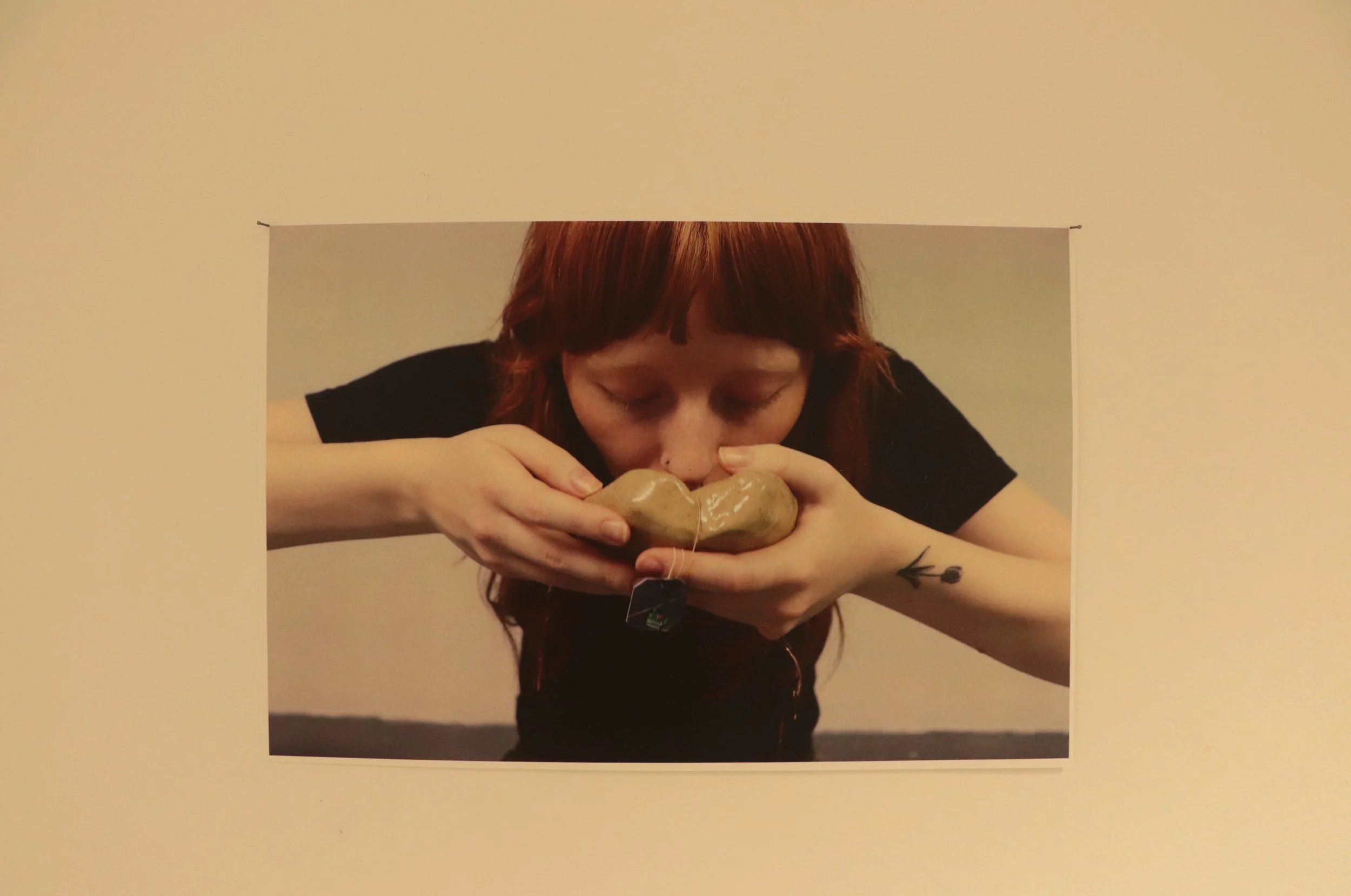 A woman with red hair and a tattoo on her left arm is holding a large, beige ceramic object near her face, with her eyes closed.