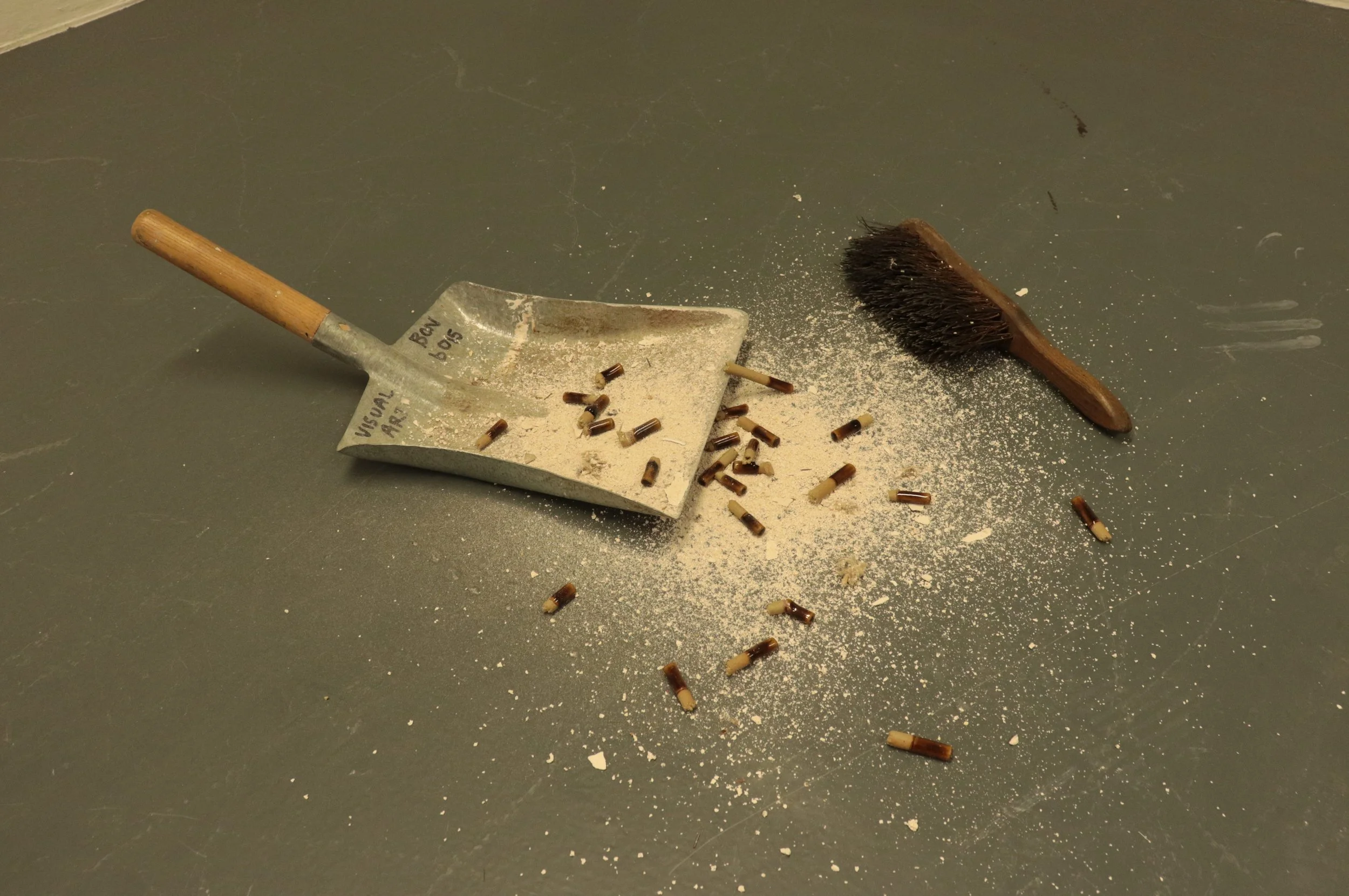 A dustpan and brush on a gray surface, with small brown and beige objects spilled over, some inside the dustpan and some on the surface.