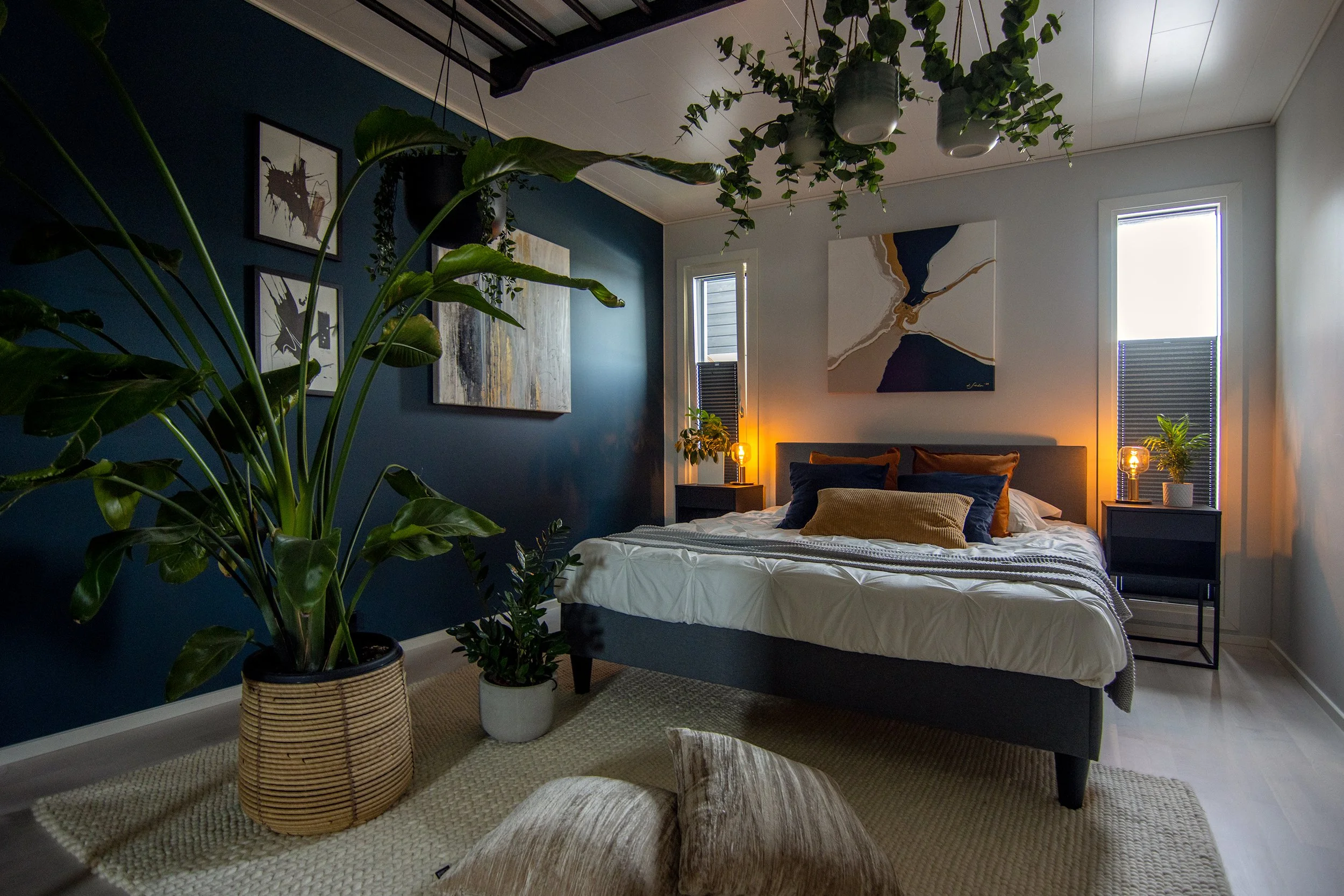 Tyylikäs makuuhuone sisustettu tumman ja vaalean sävyillä, suuret vihreät huonekasvit ja modernit taidemaalaukset seinillä.